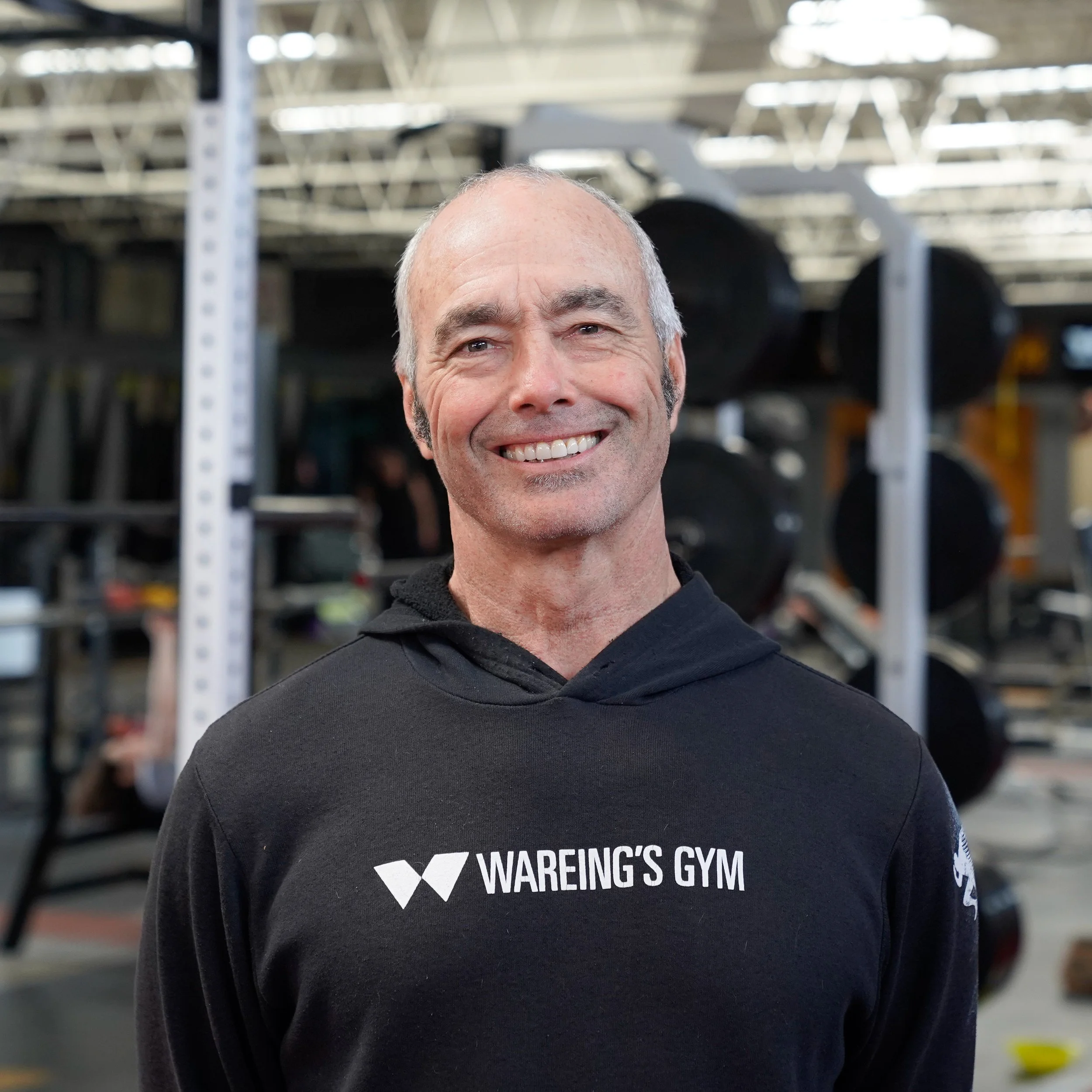 Close-up of a smiling bald man wearing a black T-shirt with 'WAREING'S GYM' logo on it, standing in a gym with shelves of gym equipment in the background.