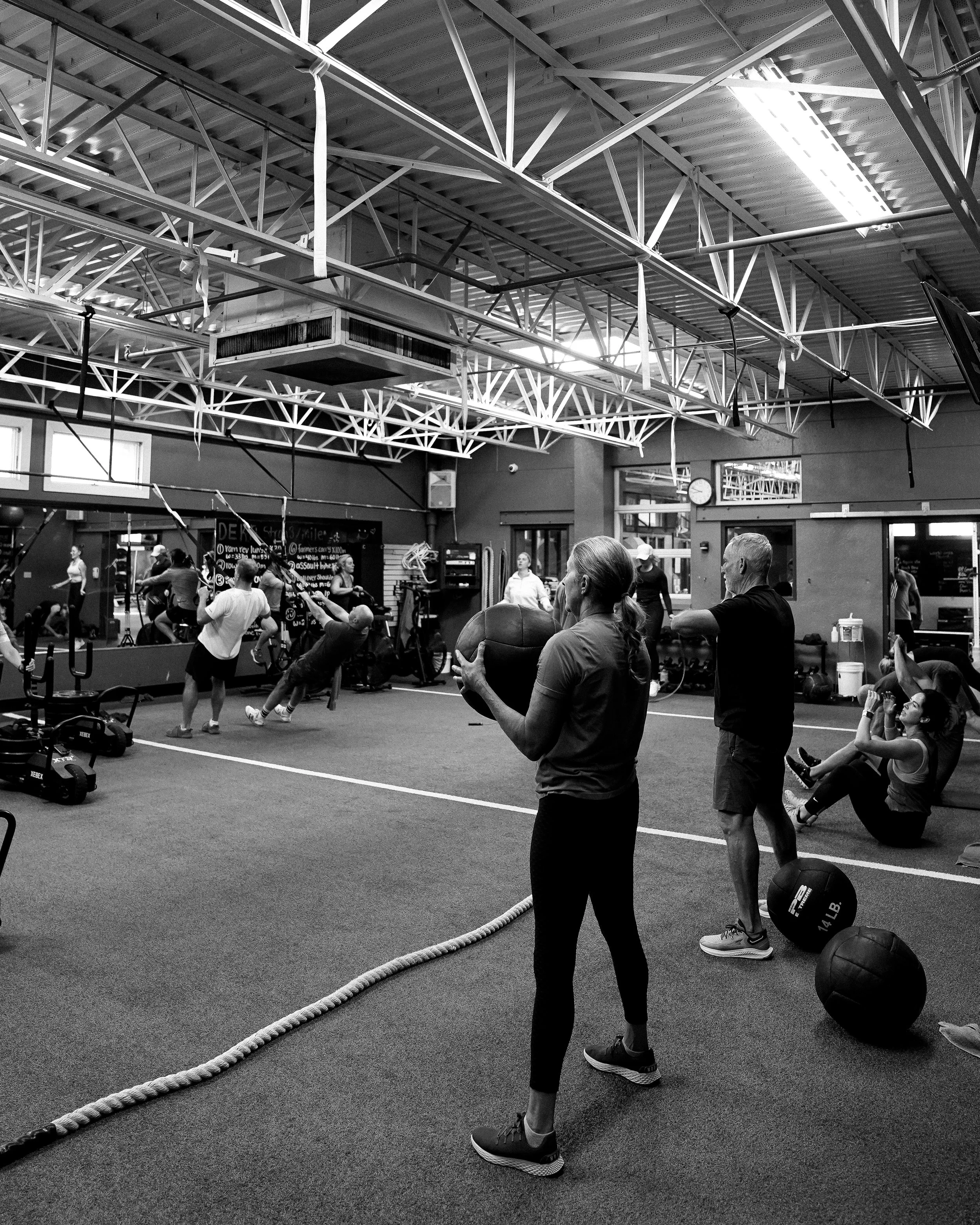 People exercising in a gym with weights, kettlebells, and ropes, under a metal ceiling with bright lighting.