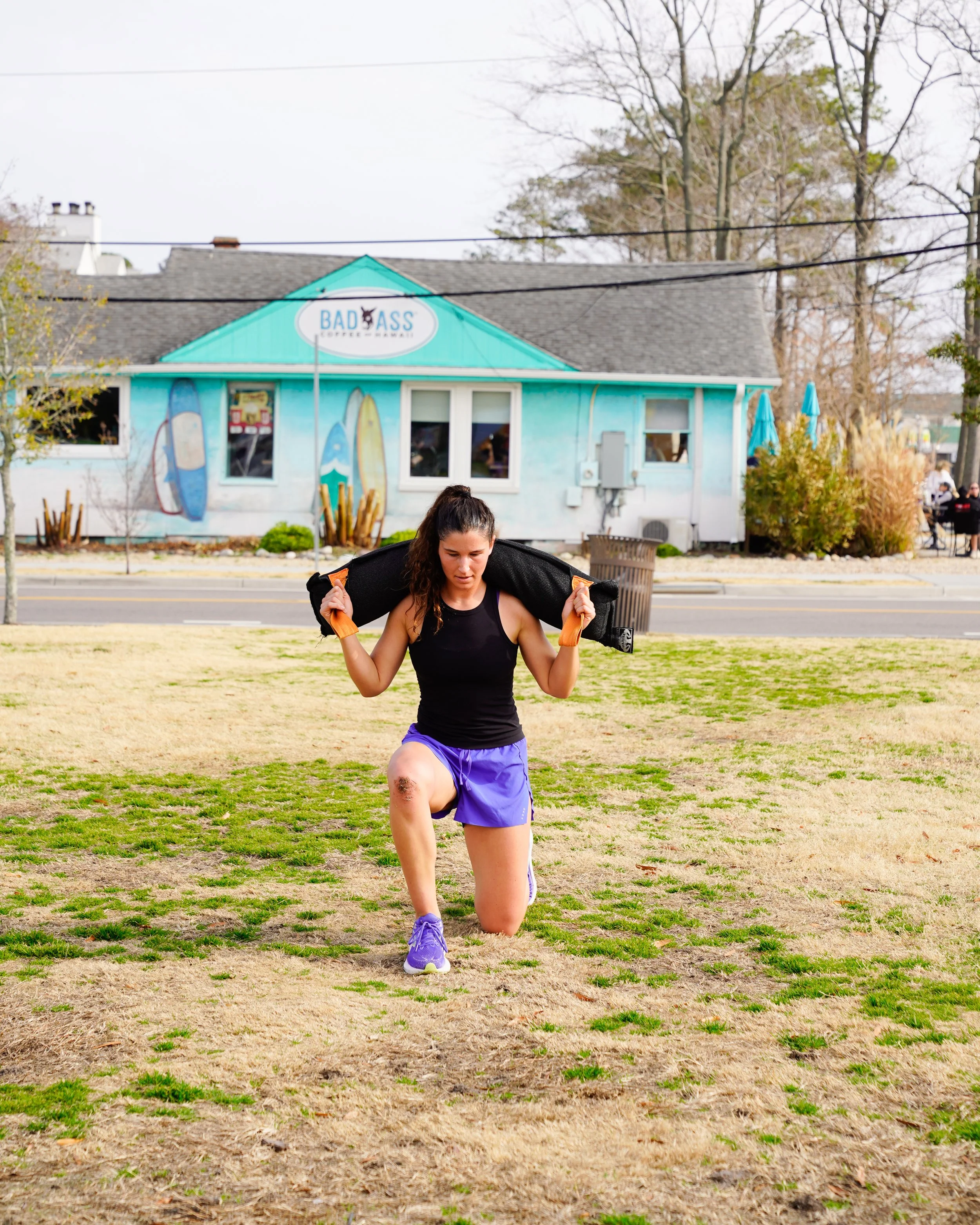 A woman wearing a black tank top and purple shorts is kneeling on one knee and holding an orange and black weighted vest behind her shoulders. She is outdoors on a patchy grass field with a building in the background that has a sign reading 'BAD ASS'