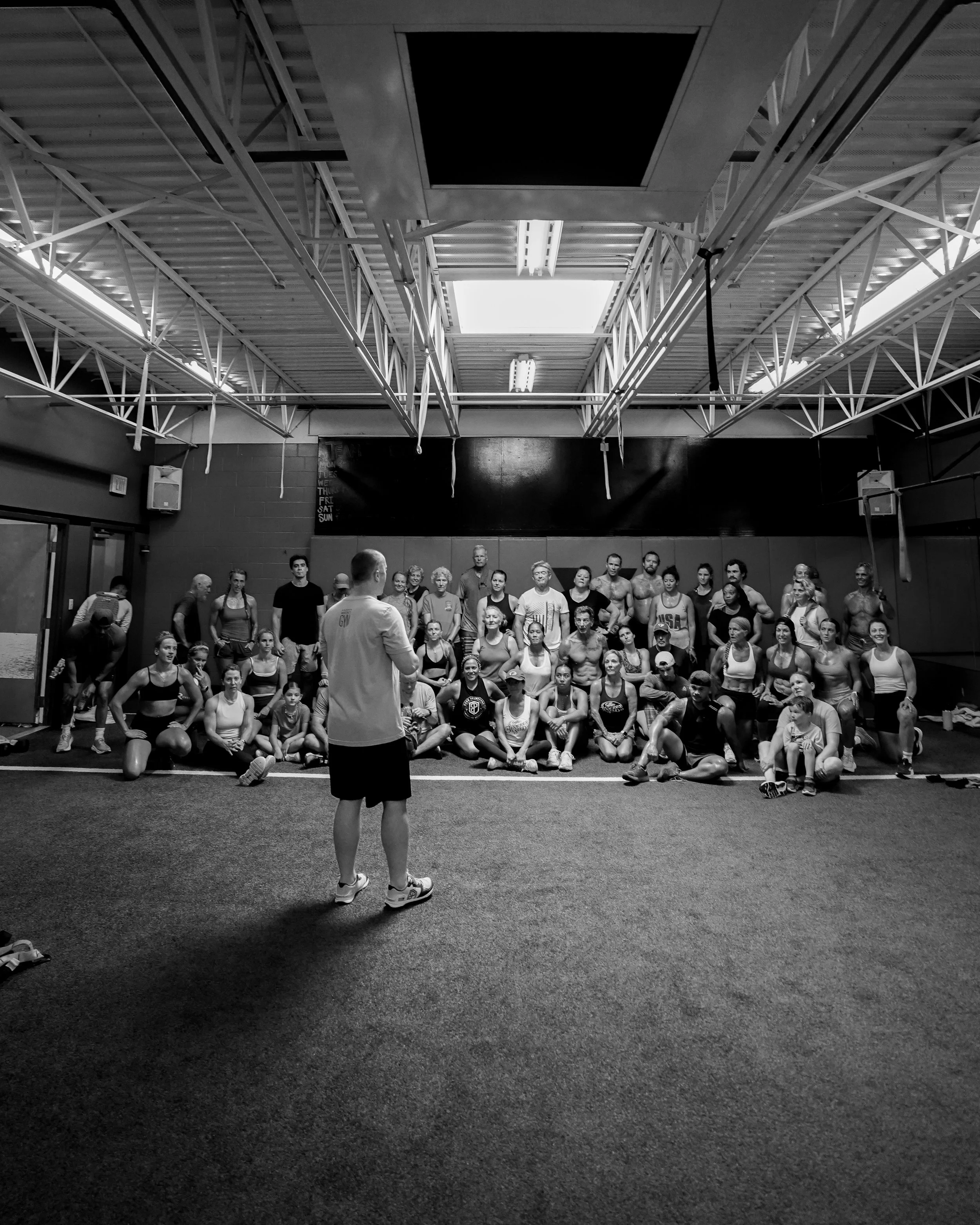 Indoor gym or fitness studio with a group of people participating in a class, some standing, some sitting, facing a male instructor. The ceiling has exposed beams and skylights, and the walls are decorated with a blackboard and some equipment.