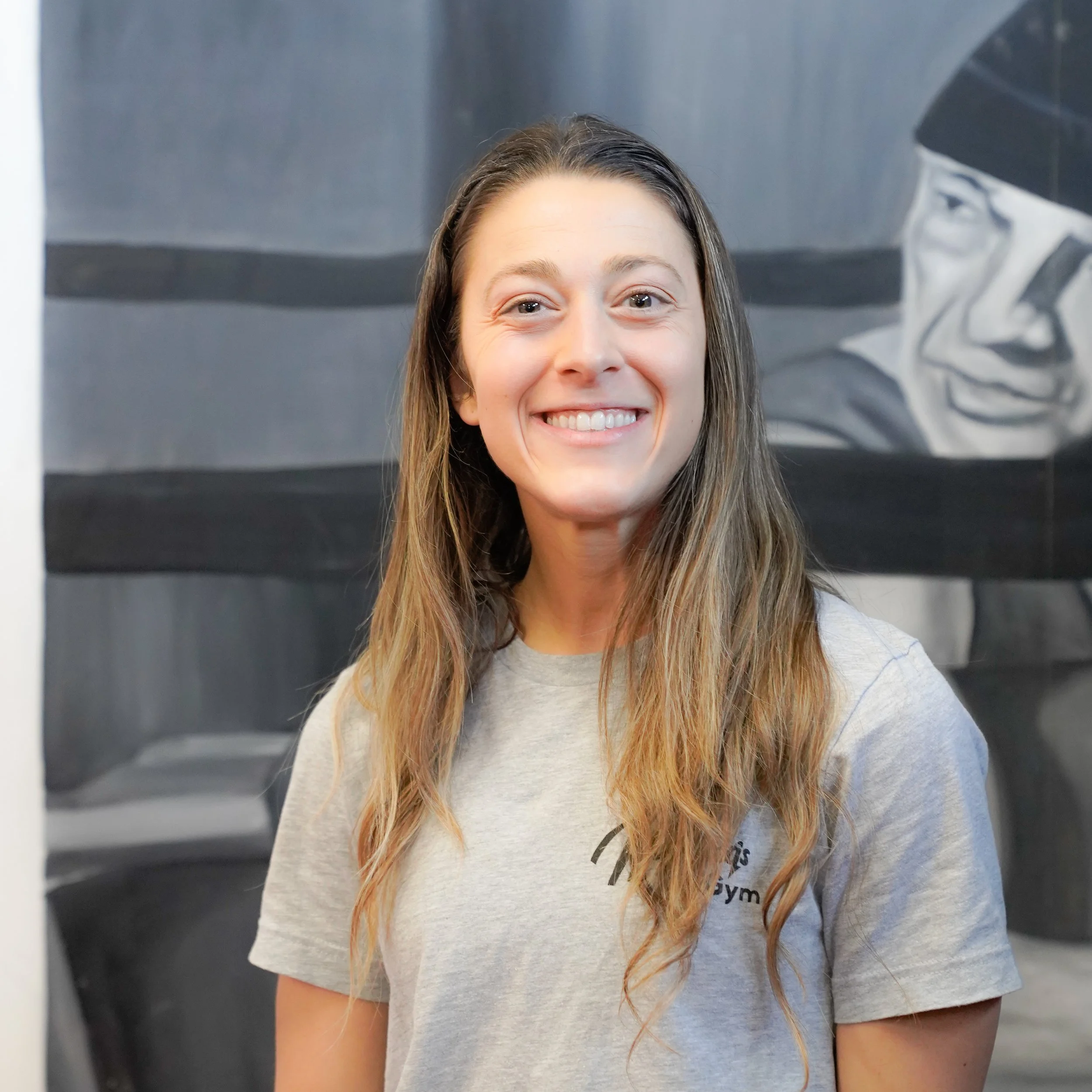 Close-up of a smiling woman with long hair wearing a grey shirt. There is a black and white portrait in the background.