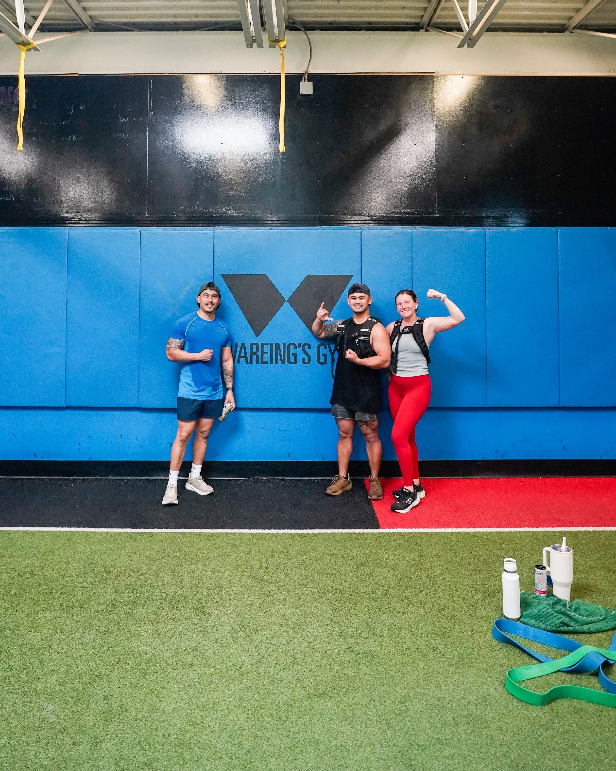 Three fitness enthusiasts standing in a gym, smiling and flexing their muscles. The gym features a black and blue padded wall with the logo and name of the gym, 'Vareing's Gym.' The ceiling has industrial exposed rafters, and the floor is wall-to-wall artificial turf with green, black, and red sections. In the foreground, there are water bottles, a basket with performance bands, and other gym accessories.