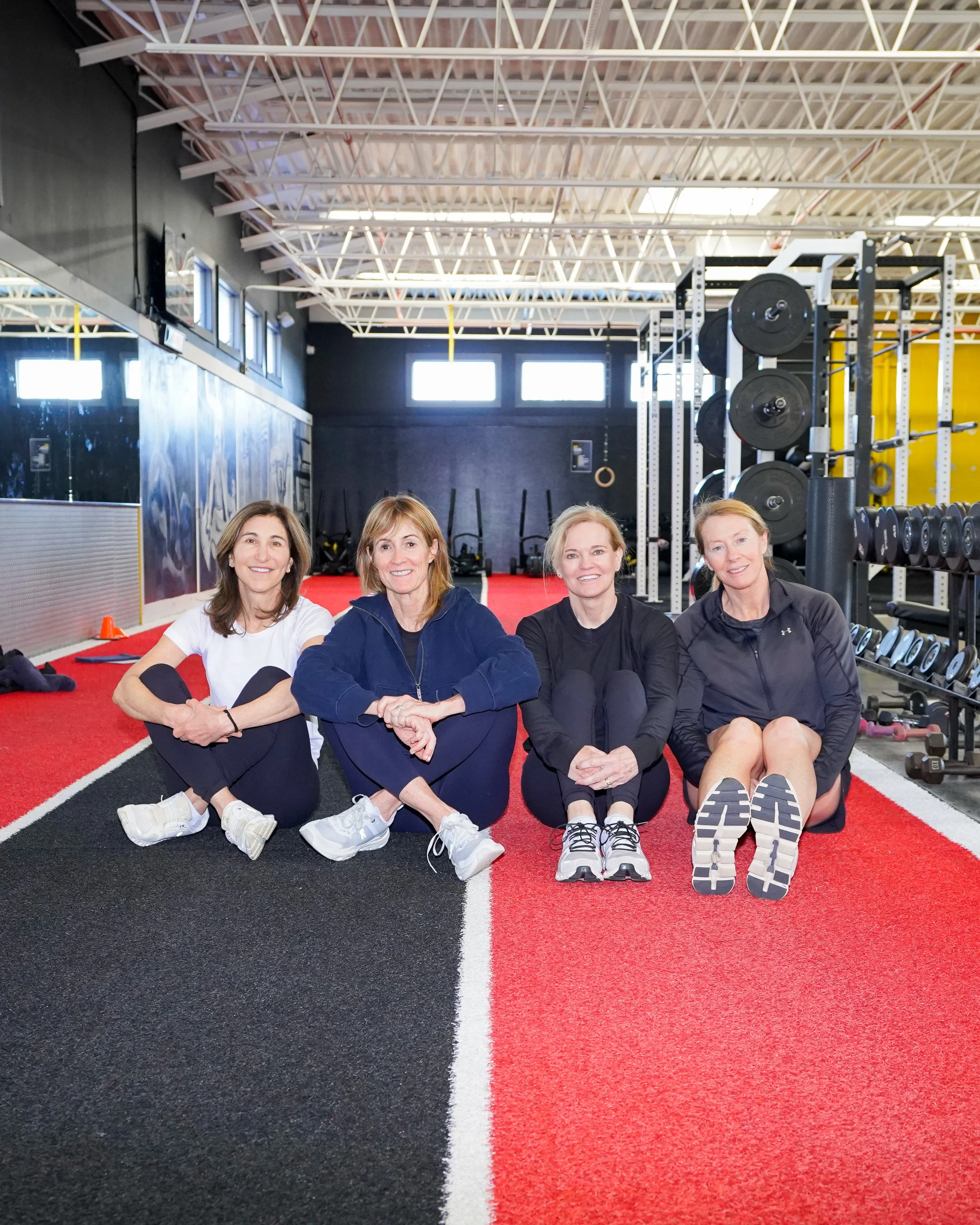 Four women sitting on a red and black running track inside a gym, posing for the photo with exercise equipment and black wall in the background.