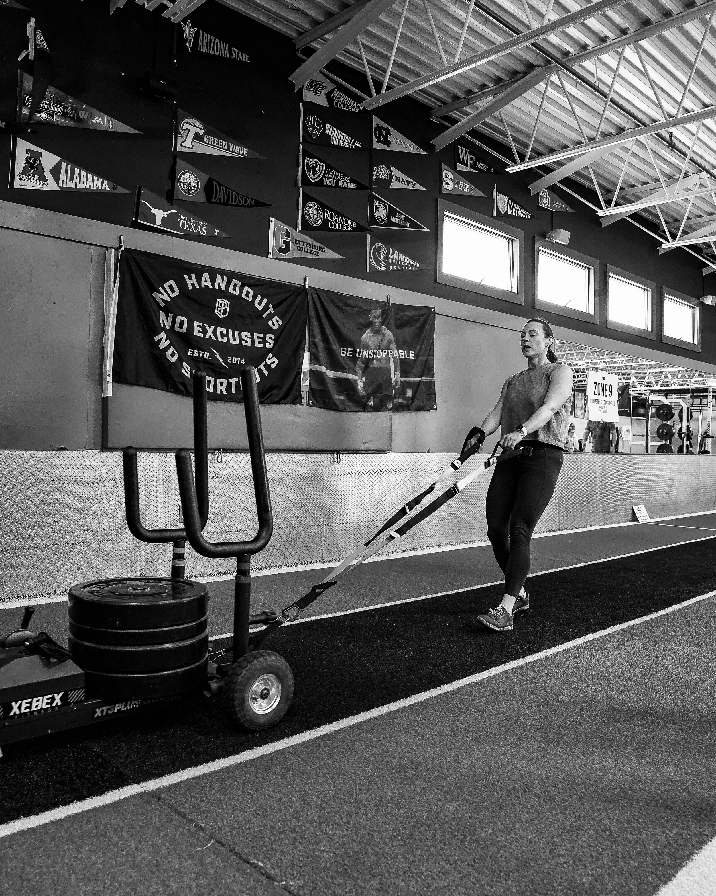 A woman using a resistance sled machine in a gym, with black and white banners of various college sports teams and flags on the wall behind her.