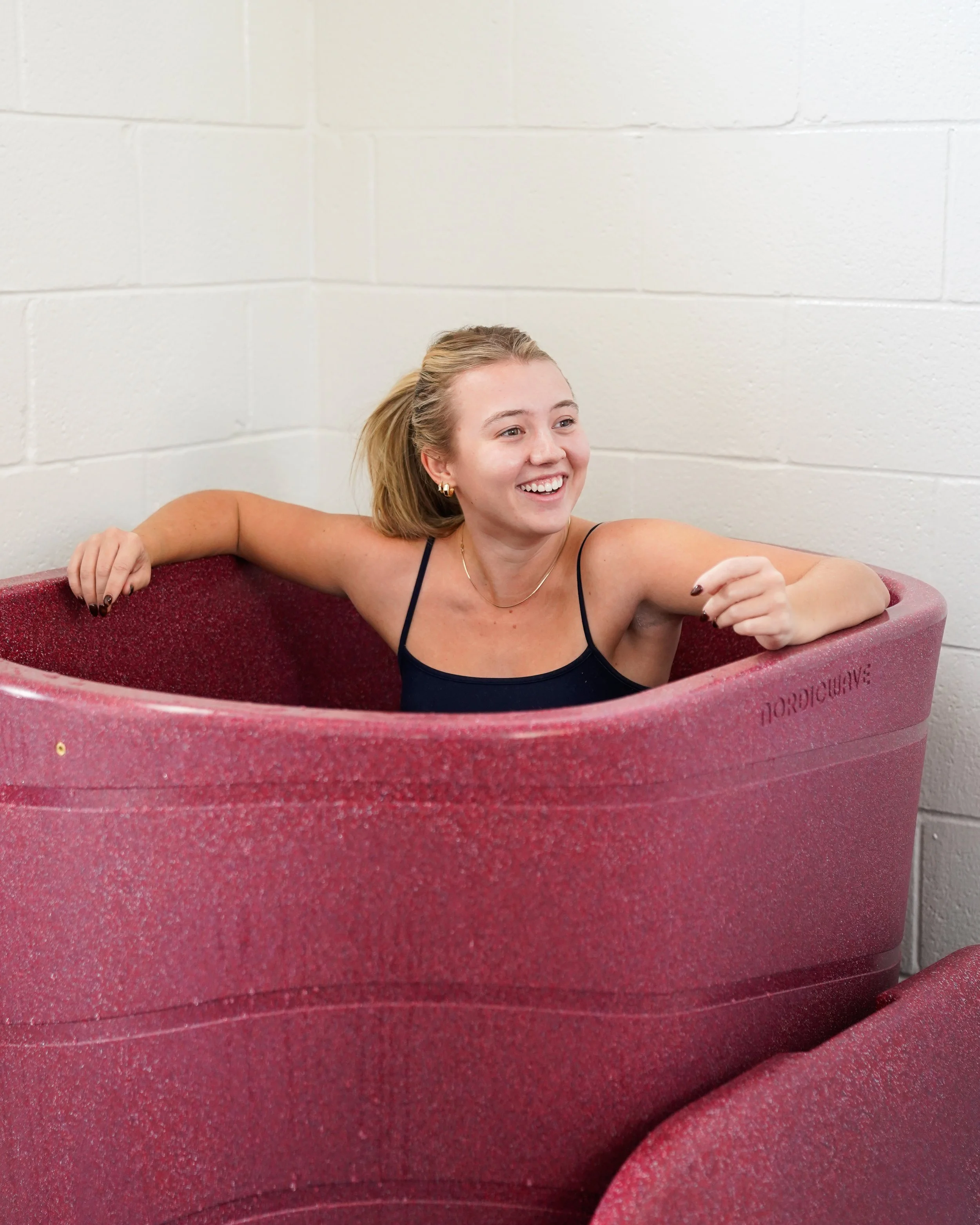 A young woman with blonde hair, wearing earrings and a black spaghetti strap top, is smiling and relaxing in a round, pink, textured bathtub with her arm resting on the edge, against a plain white brick wall.