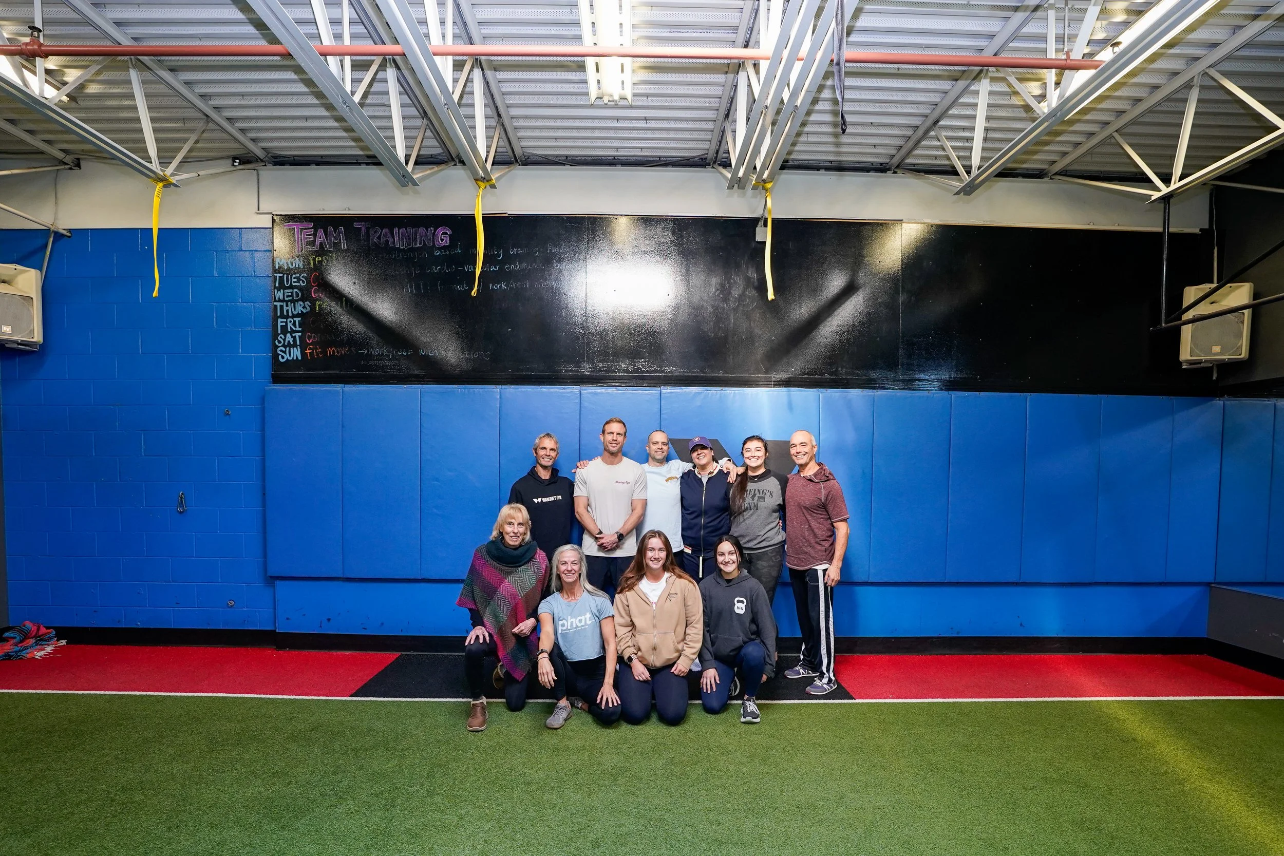 Group of ten people, consisting of adults and children, standing and kneeling on a green and red artificial turf in a gym or sports facility, posing for a photo with a blackboard and gym equipment in the background.