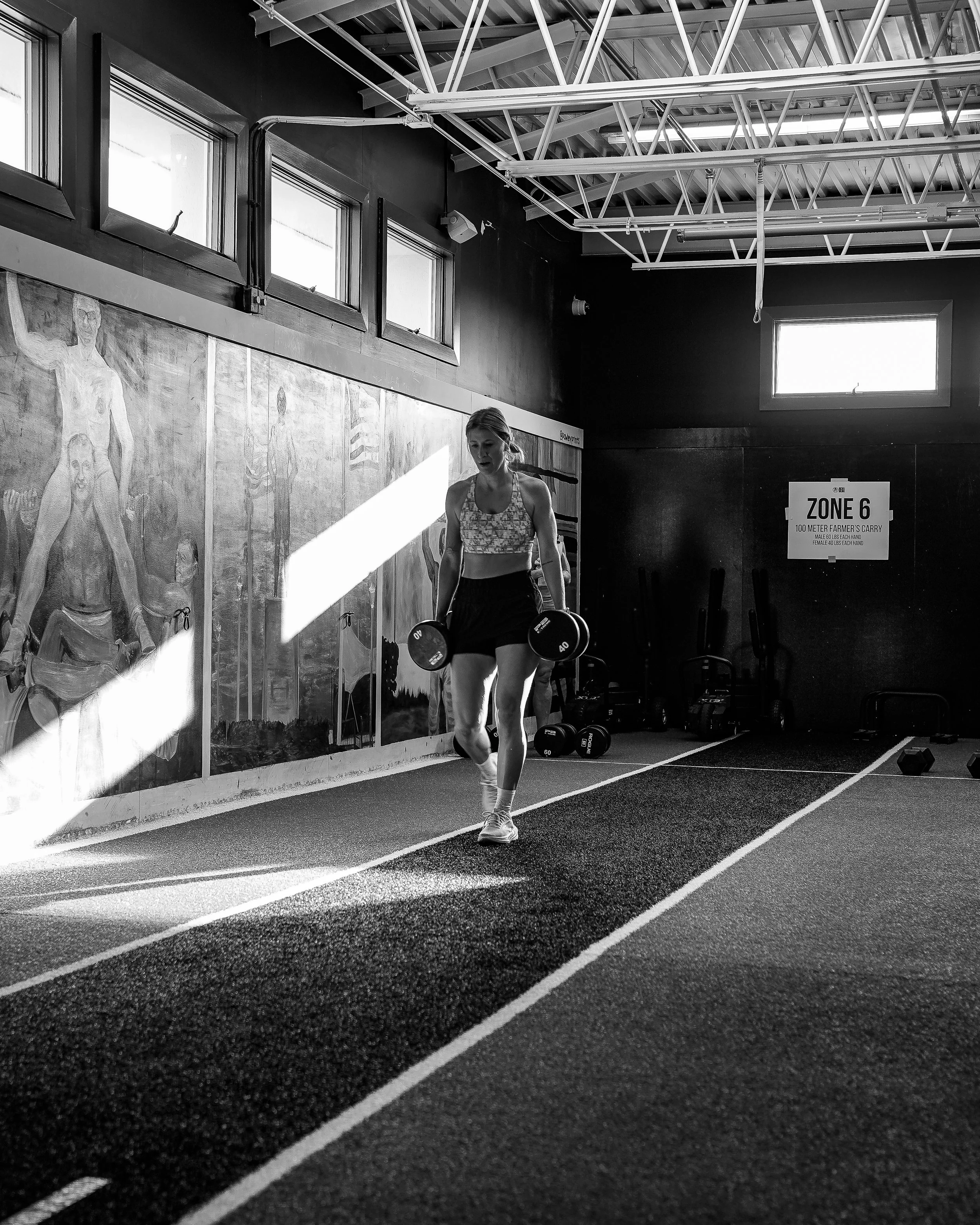 A woman lifting a dumbbell while walking on a treadmill inside a gym with a mural wall and a sign that reads 'Zone 6, 100 Meter Farmer's Carry'.