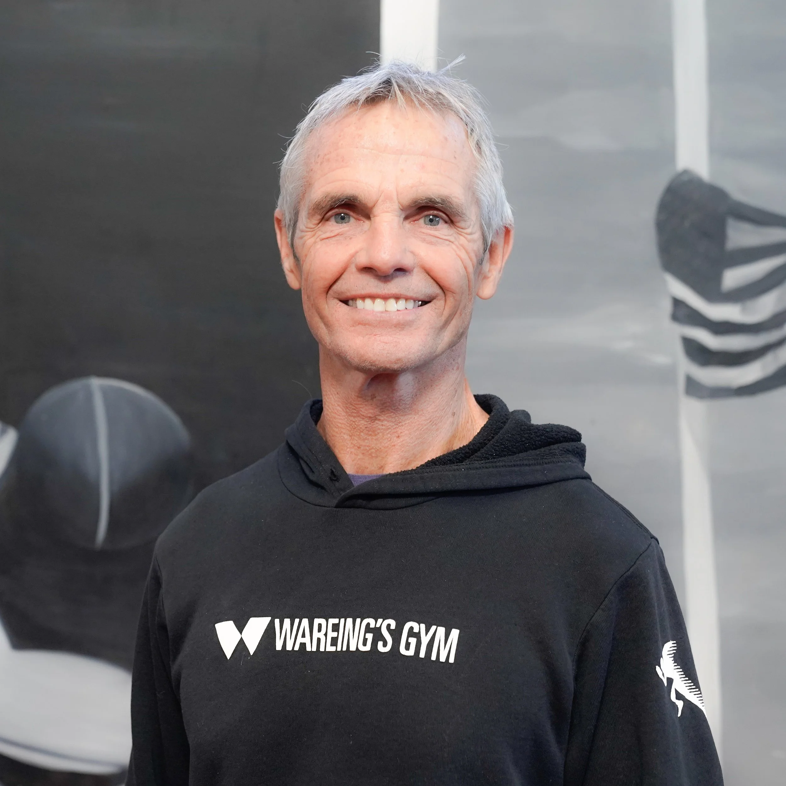 Close-up of a smiling older man with gray hair wearing a black hoodie with 'WAREING'S GYM' printed on it, standing in front of a dark background with a logo.