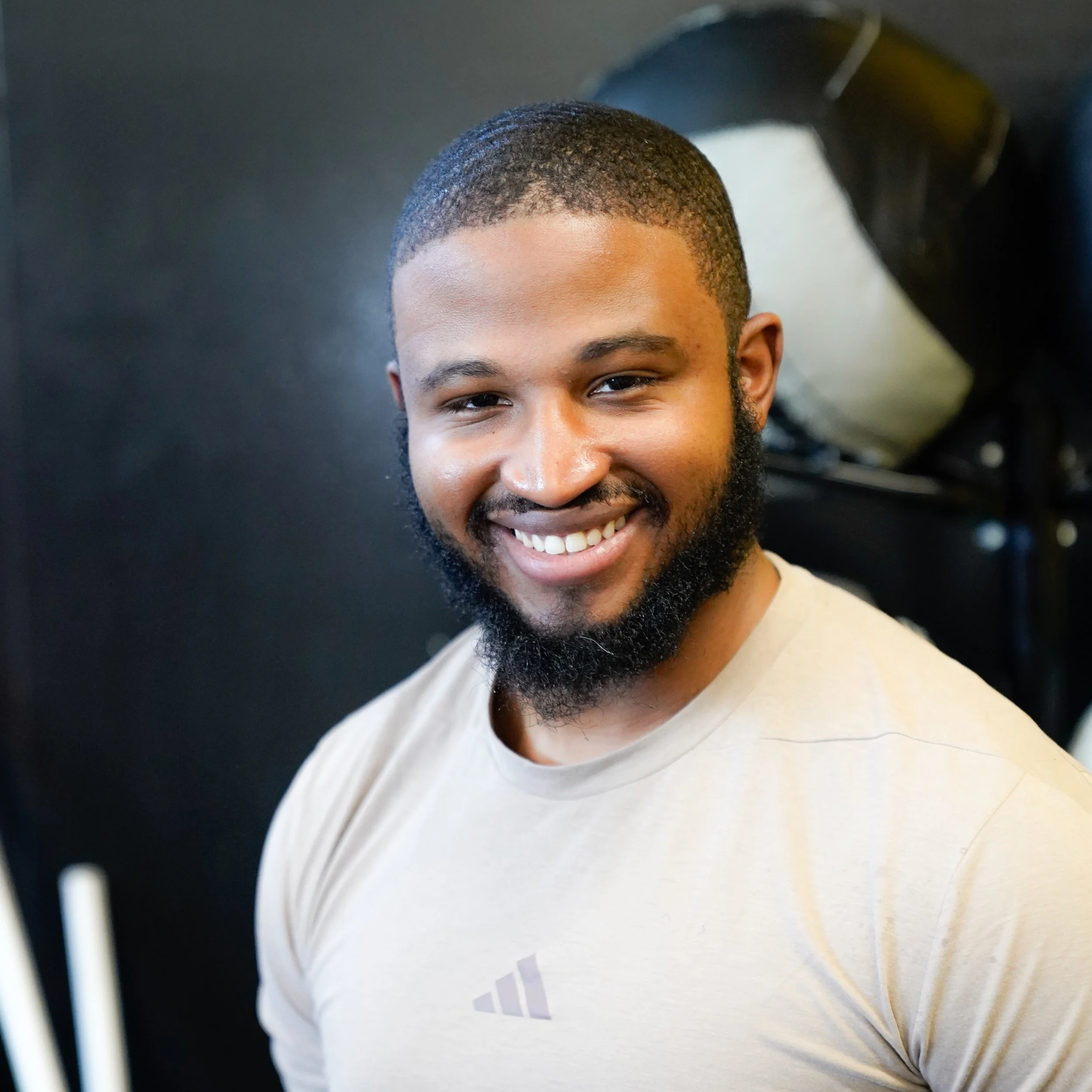 Close-up of a young man with short curly hair and a beard smiling, wearing a beige shirt, against a blurred indoor background.