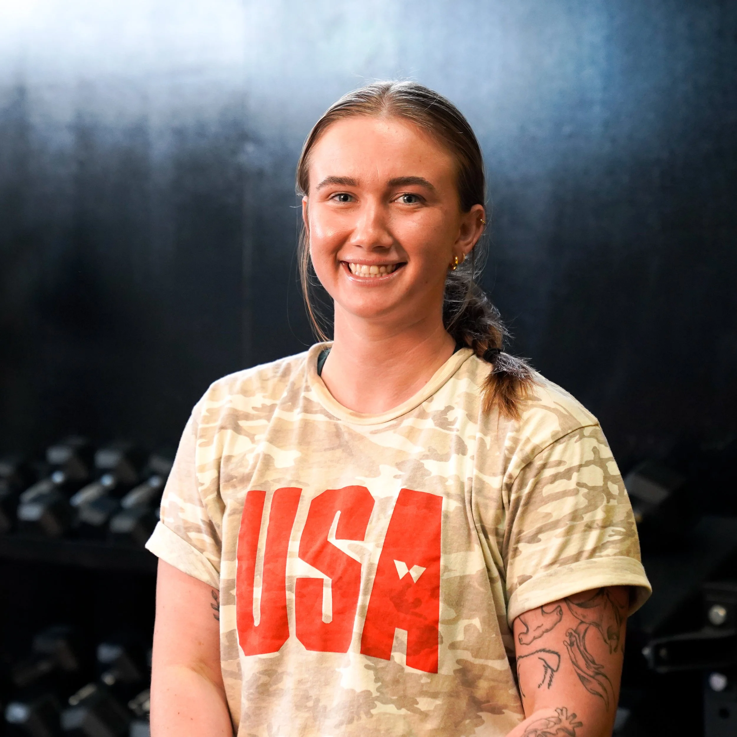 A young woman with light skin and long brown hair tied back, smiling, wearing a beige camouflage T-shirt with red text, and gold hoop earrings against a dark background.