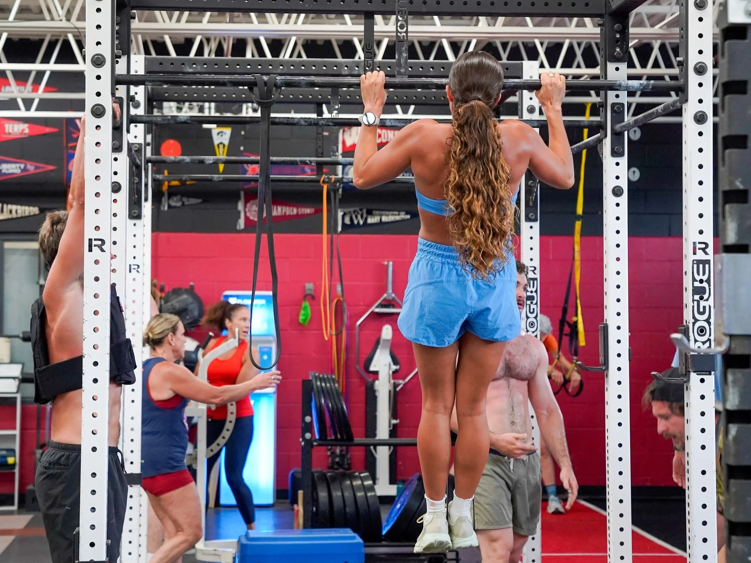 A woman with long curly hair, wearing a blue sports bra and shorts, is performing pull-ups on a gym rack. Several other people are in the background, some measuring weights and others sitting or standing nearby. The gym has a red and black theme with various equipment around.