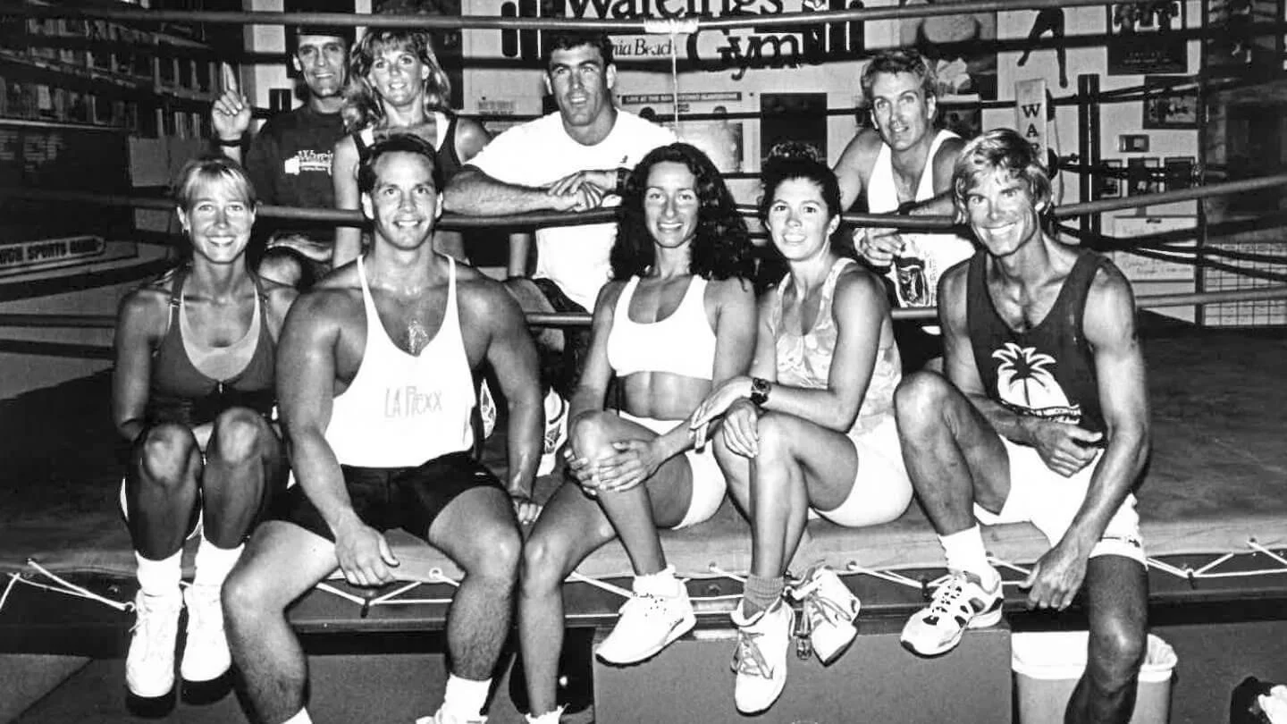 Black and white photo of a group of nine people, six men and three women, sitting and standing in a gym with boxing or wrestling ring ropes in the background. They are dressed in athletic workout attire, smiling and posing for the camera.