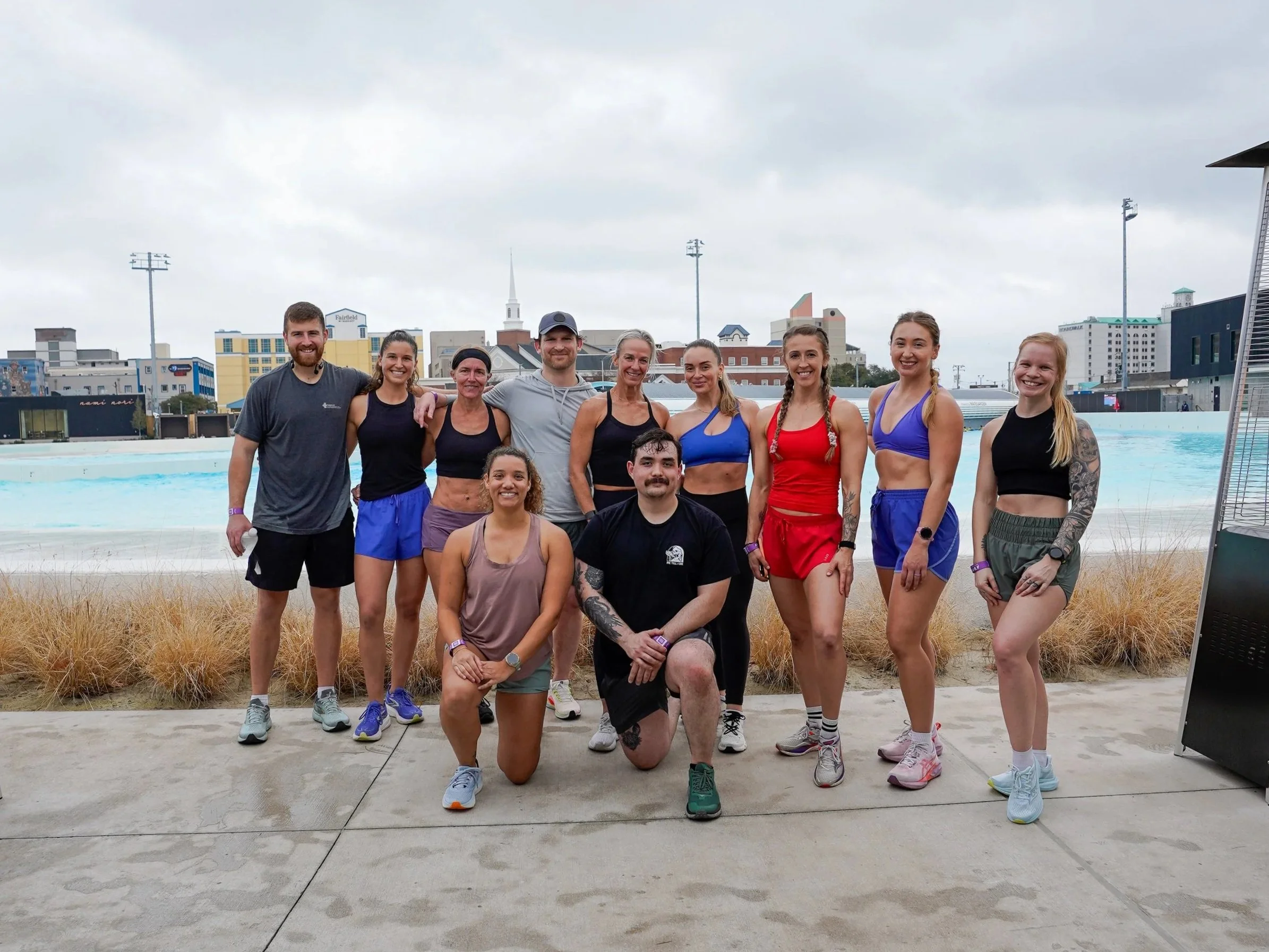 Group of ten people, fitness enthusiasts, posing together outdoors by or in a shallow pool in a city with modern buildings, cloudy sky, some in athletic wear, smiling at camera.