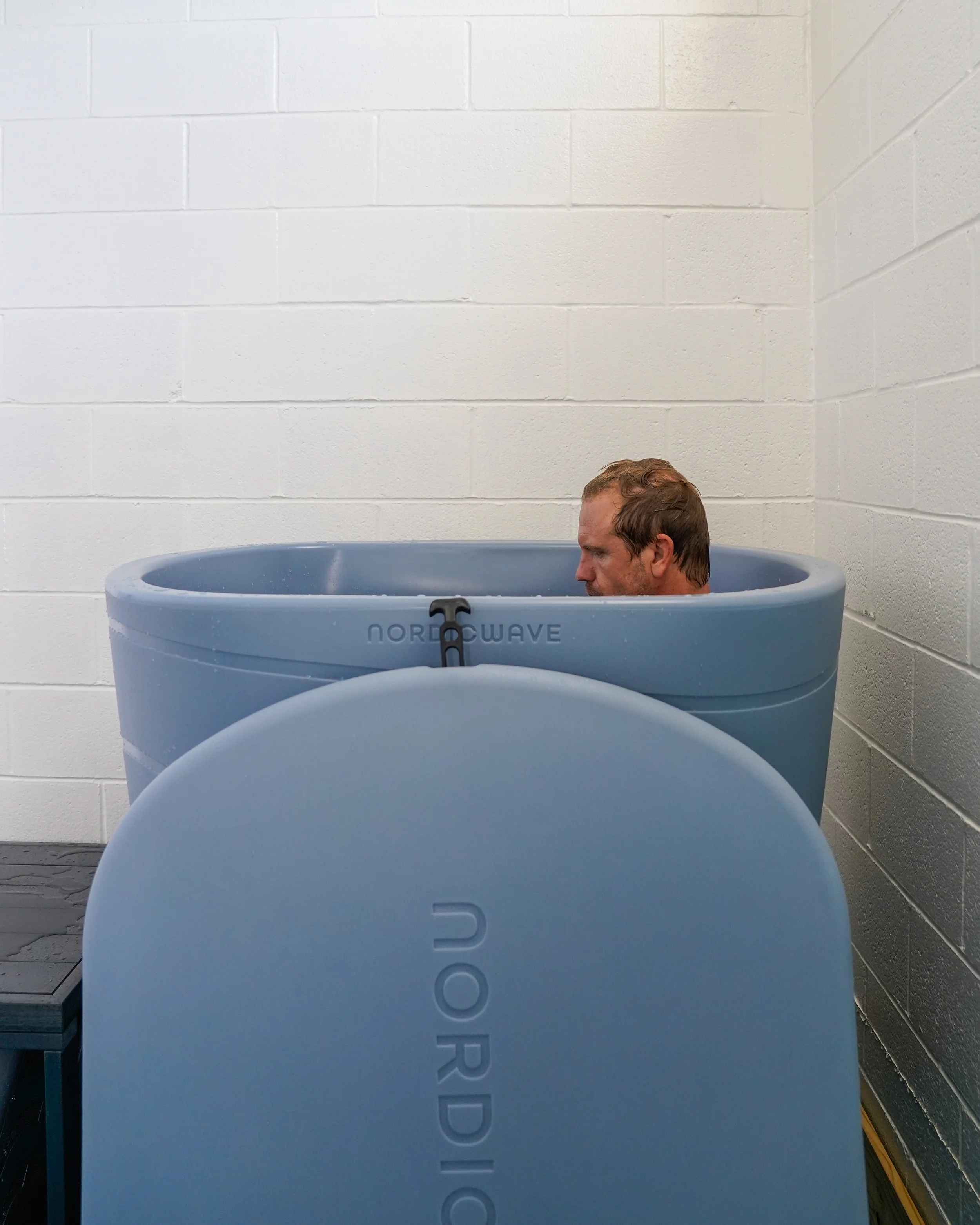 A man with short brown hair and light skin sitting in a blue NordIce IceBath tub against a white brick wall.