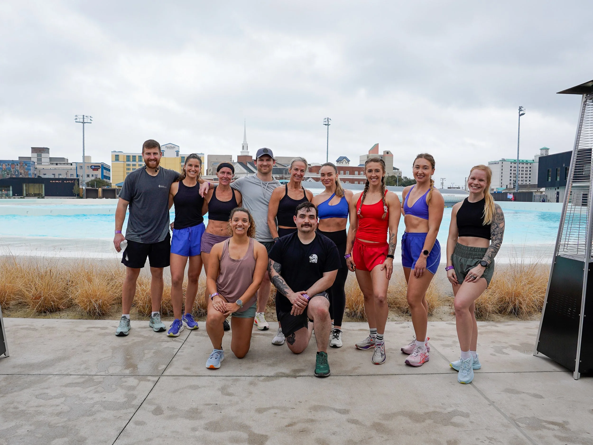 A group of eleven fitness enthusiasts posing outdoors near a swimming pool in an urban area, with city buildings and a cloudy sky in the background.