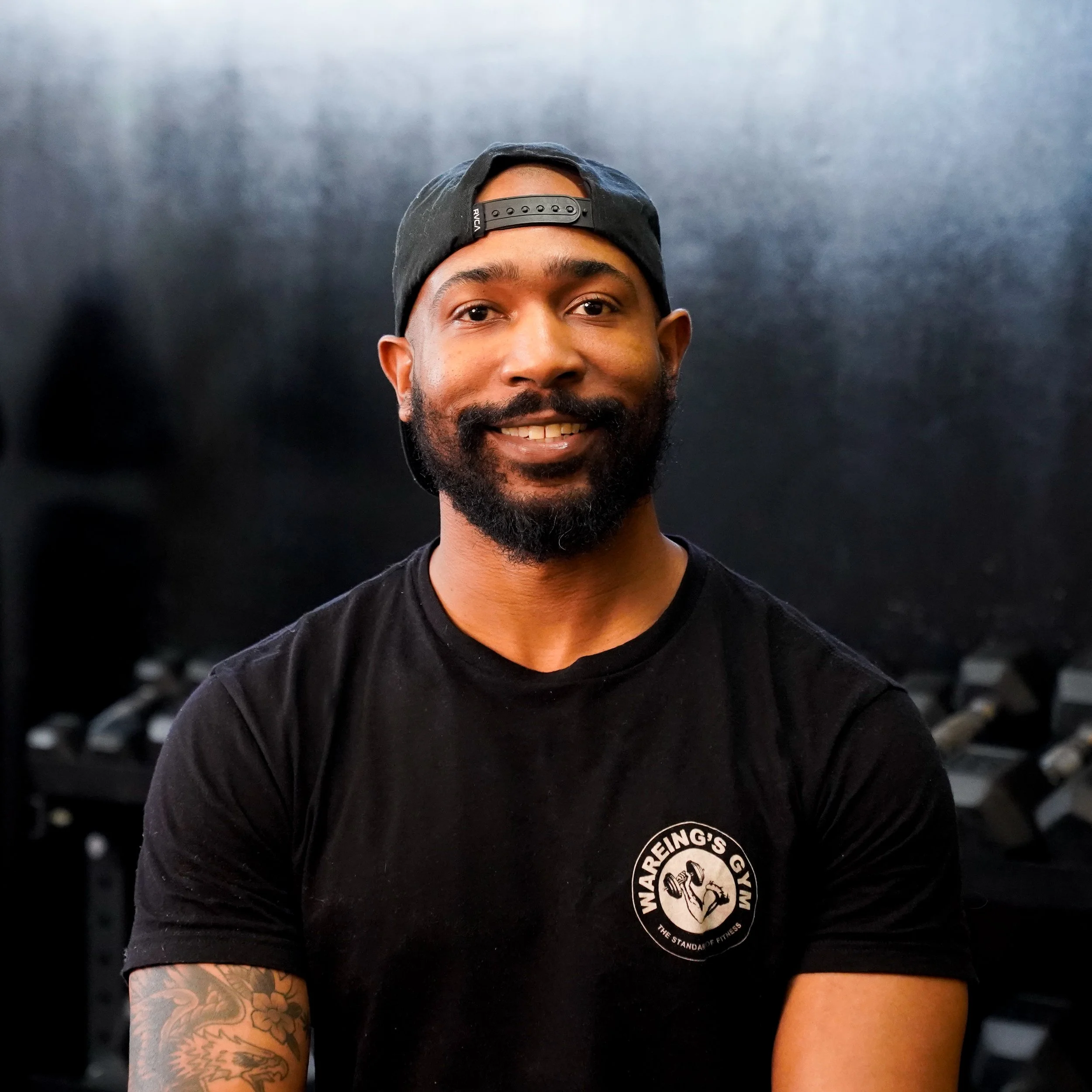 Close-up of a young man with a beard wearing a black baseball cap backwards and a black T-shirt.