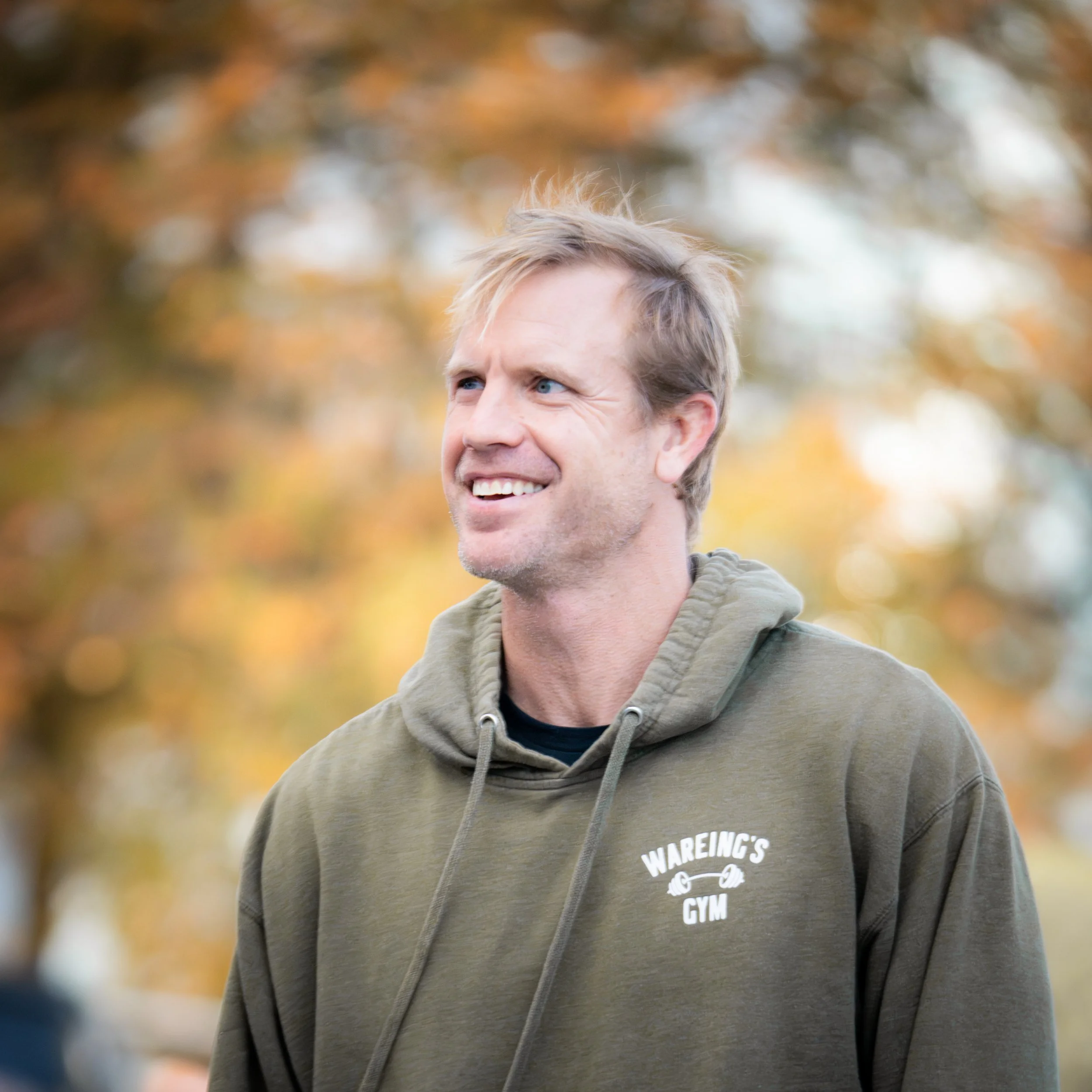 Close-up of a smiling man with blonde hair and blue eyes wearing a gray hoodie with the text 'WAREING'S GYM' and a barbell logo, outdoors with blurred autumn leaves in the background.