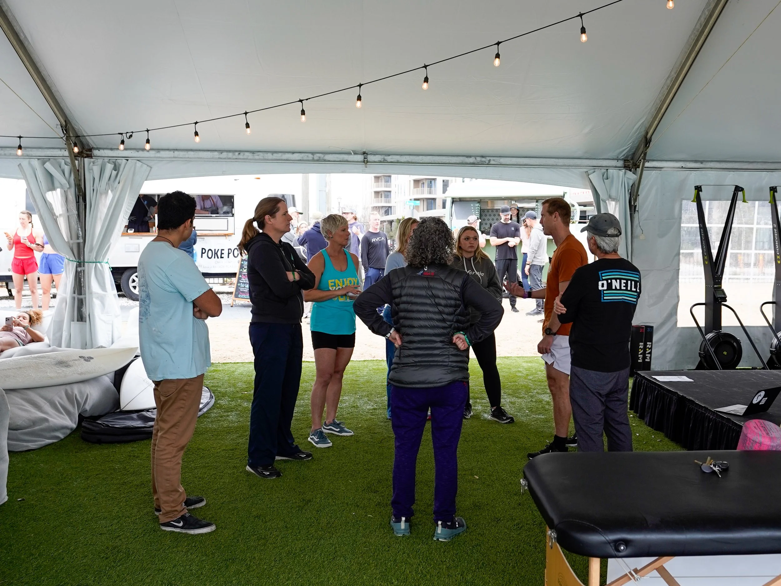 People gathered inside a large white tent outdoors, engaged in conversation. The tent has string lights hanging from its ceiling, and outside are background buildings and people, some in athletic attire.