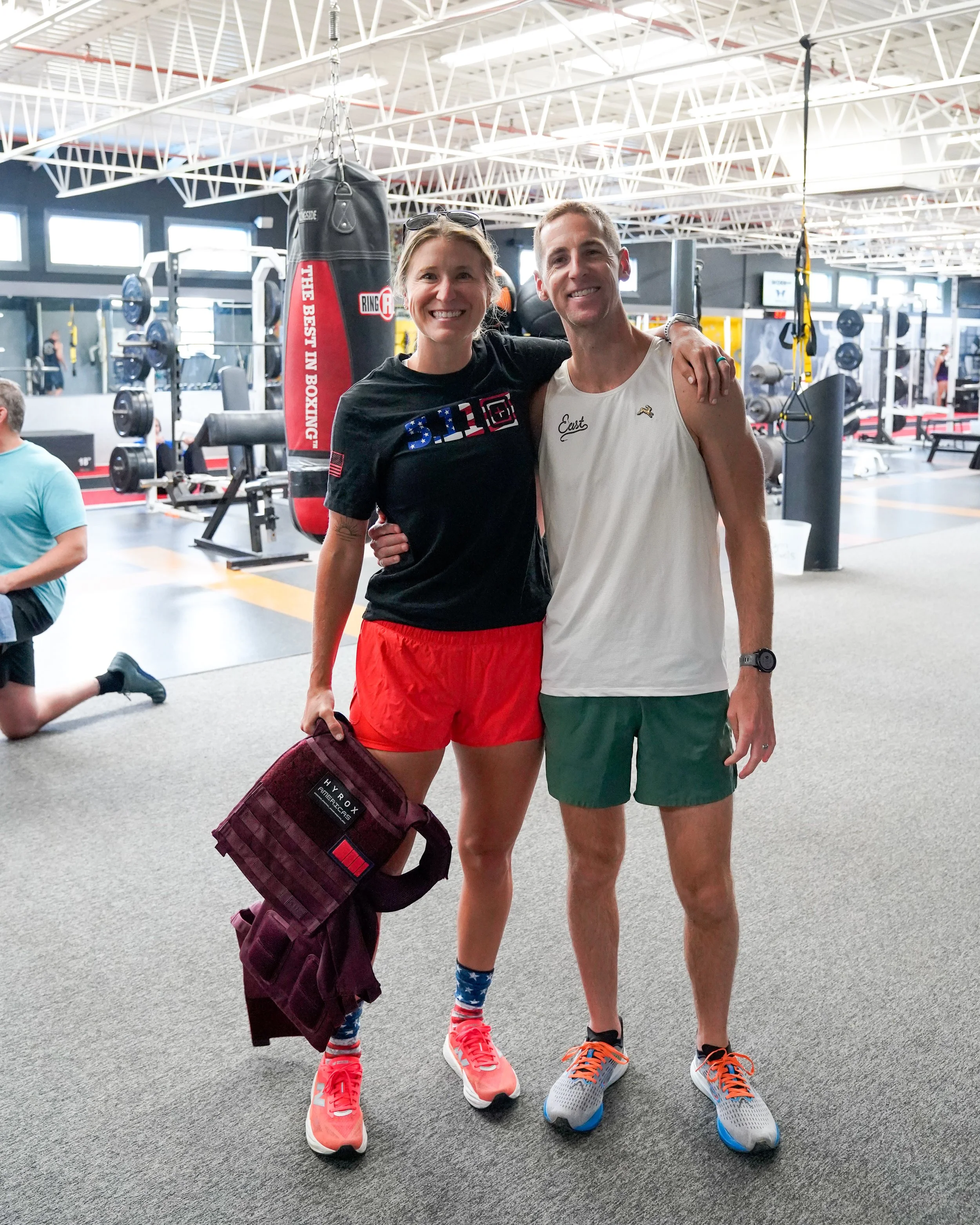 A smiling woman and man standing together inside a gym, with their arms around each other. The woman is holding a maroon backpack with a gym brand label. They are wearing athletic clothing, and the gym has workout equipment and a hanging punching bag