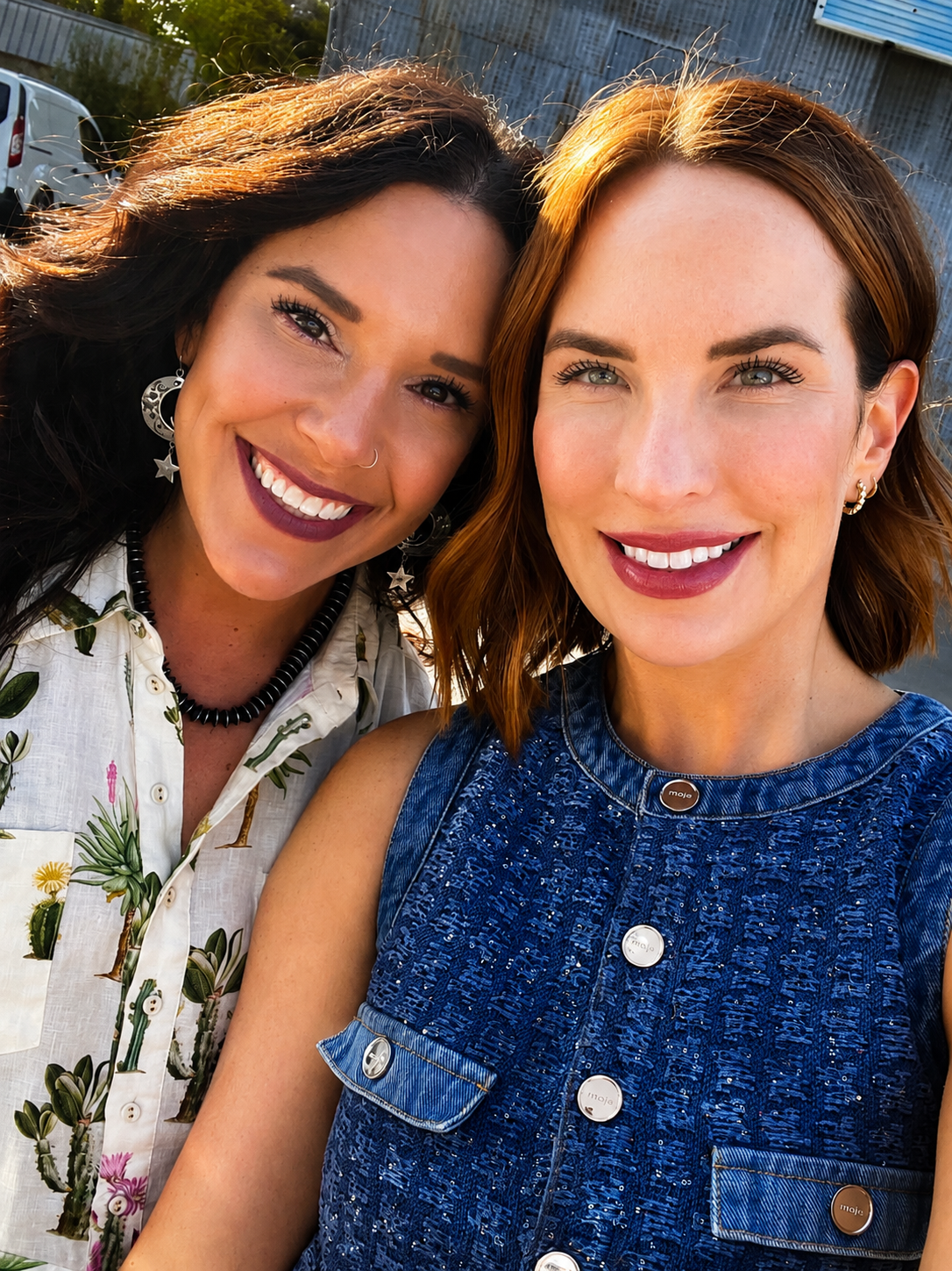 Two women smiling closely for a selfie outdoors, with cars and a building in the background.