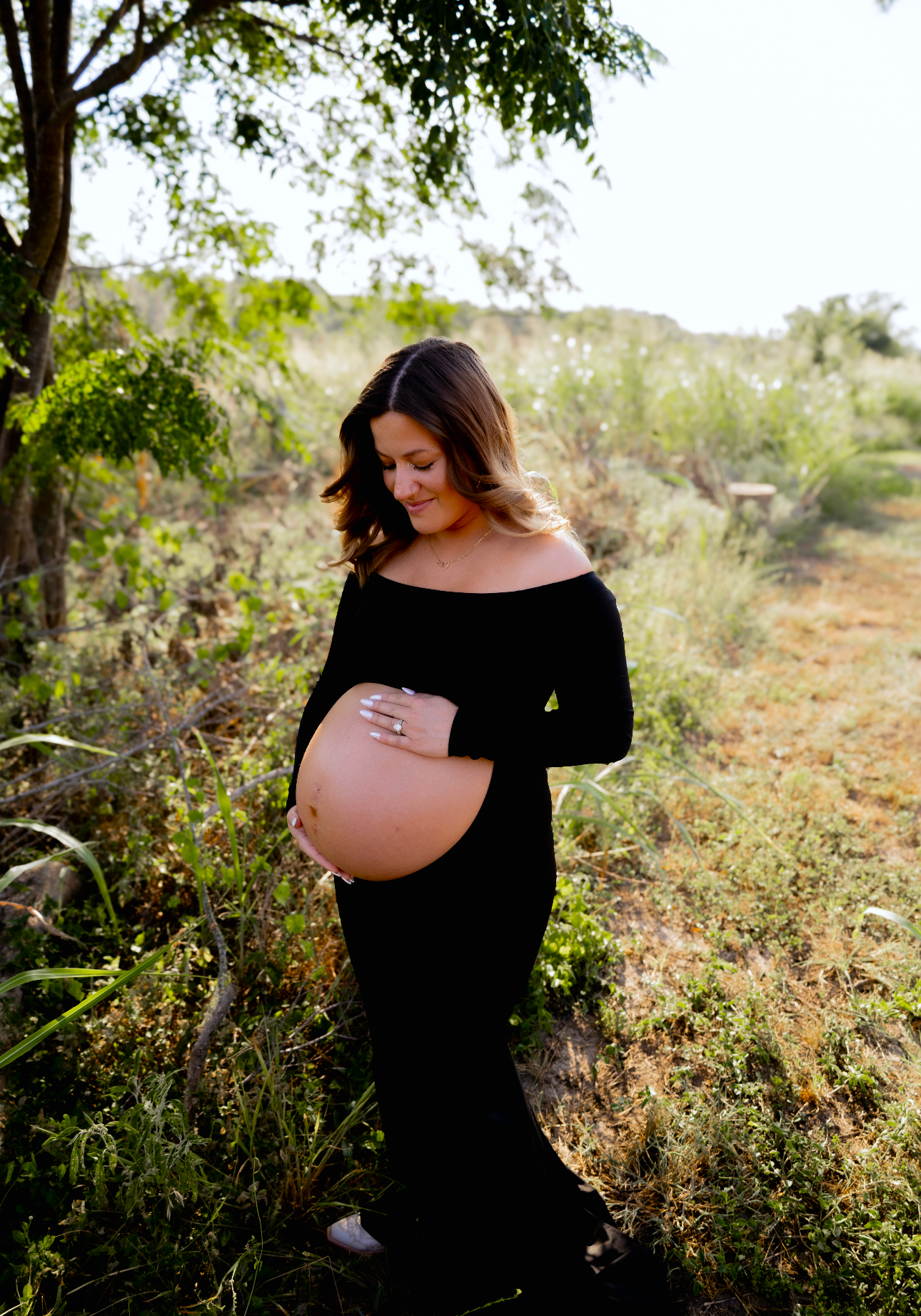 Pregnant woman in a black off-shoulder dress with wavy shoulder-length hair, standing outdoors in a lush green field, gently cradling her belly and smiling with eyes closed.