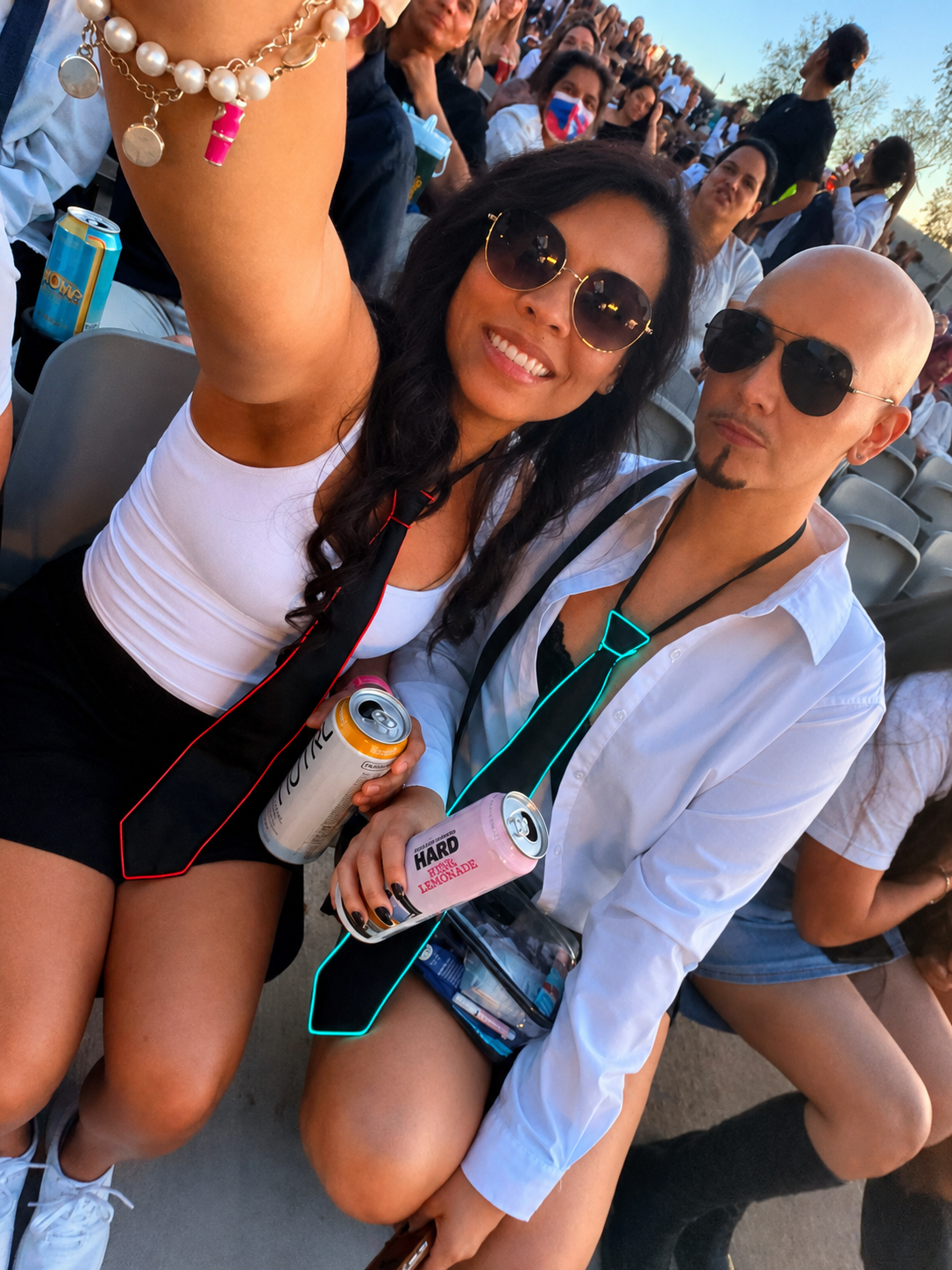 Two people sitting in an outdoor event, wearing sunglasses and holding canned drinks, smiling for a selfie. The woman has long dark hair, and the man is bald with a goatee. Many people are in the background.