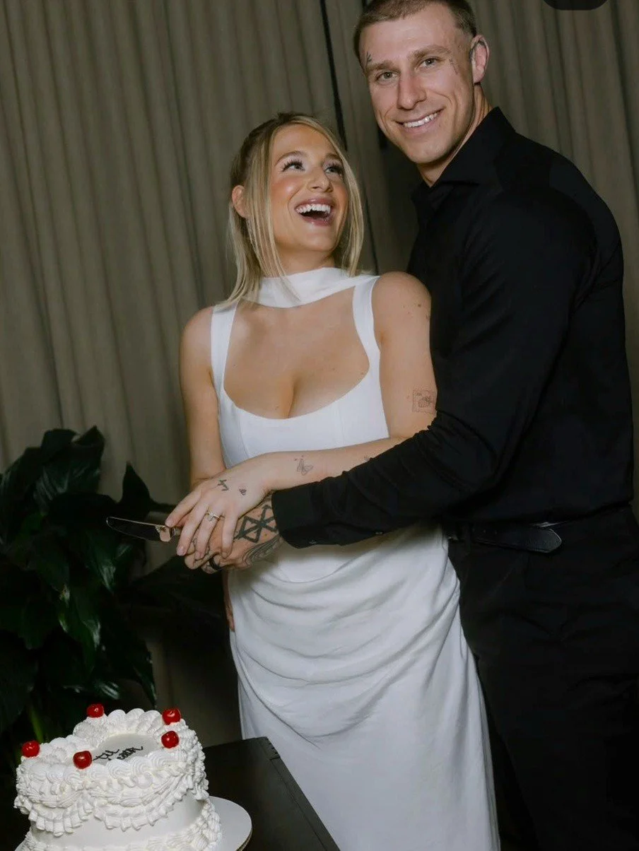 A woman in a white dress and a man in a black shirt smiling and posing together at a celebration. There is a white frosted cake with cherries on top in front of them.