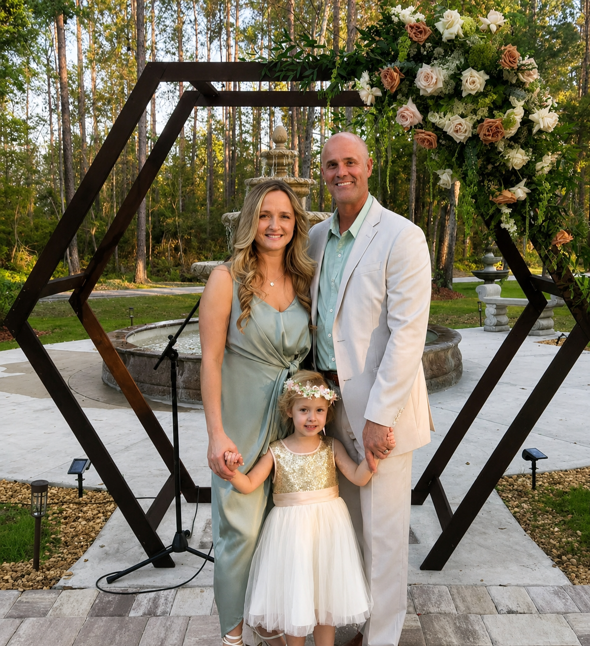 A family of three, standing hand-in-hand in front of a fountain and floral arch at an outdoor wedding venue. The woman is wearing a light satin dress, the man is in a white suit, and a young girl in a dress with a floral headband.