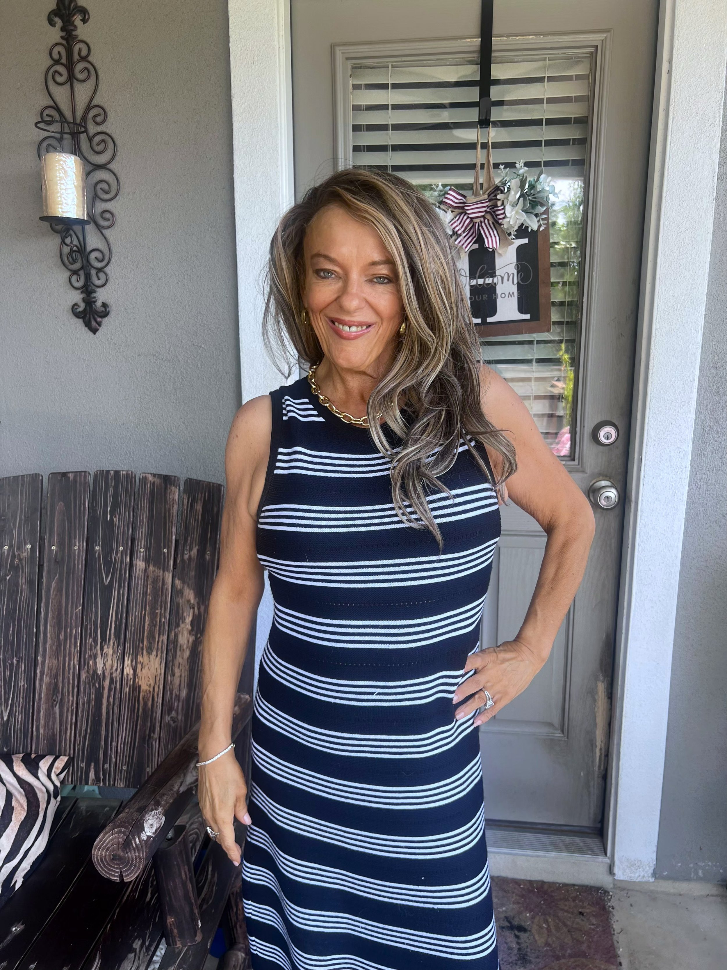 A smiling woman with long wavy hair wearing a navy and white striped dress, standing outside next to a door decorated with a sign and a bow, with a wooden bench nearby.