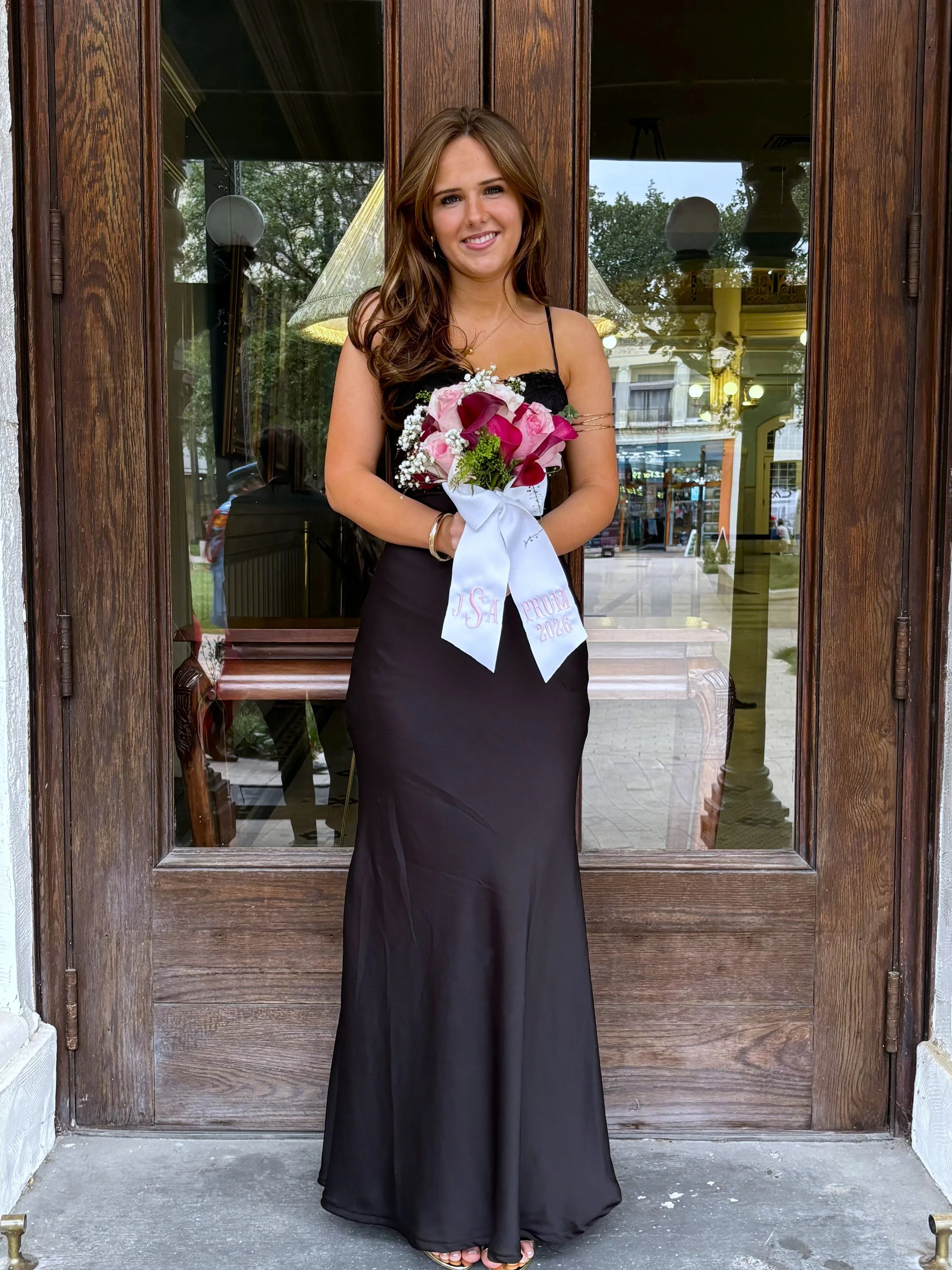 A young woman in a black dress holding a bouquet of pink and white flowers, standing in front of a wooden door with glass windows.