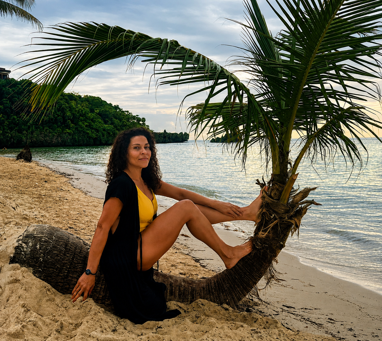 A woman sitting on a bent palm tree trunk on a beach near the ocean, with green trees in the background and a partly cloudy sky.
