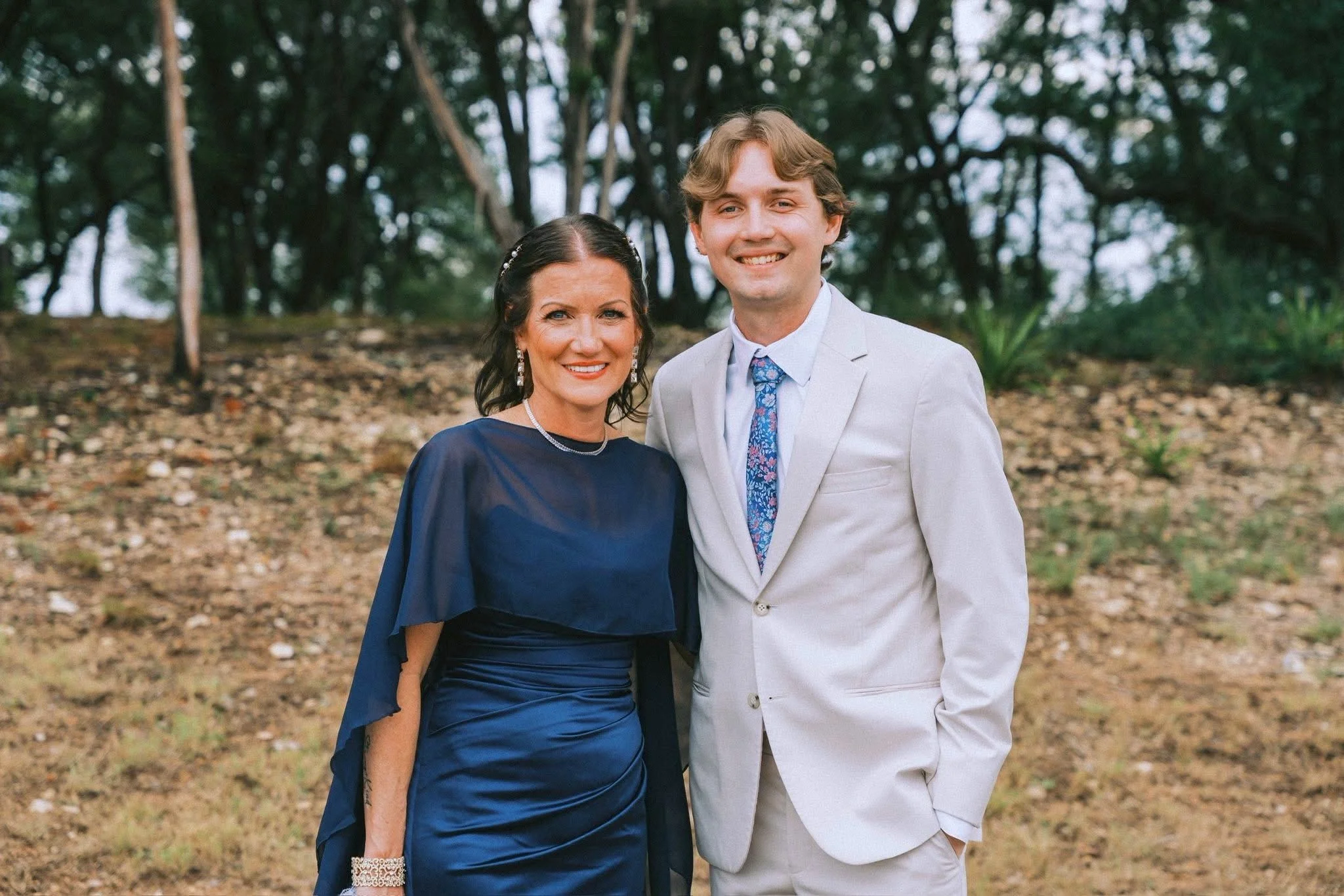 A woman in a blue dress with earrings and a necklace, and a man in a light gray suit with a floral tie, are smiling and standing close together outdoors in front of trees and a forest floor.