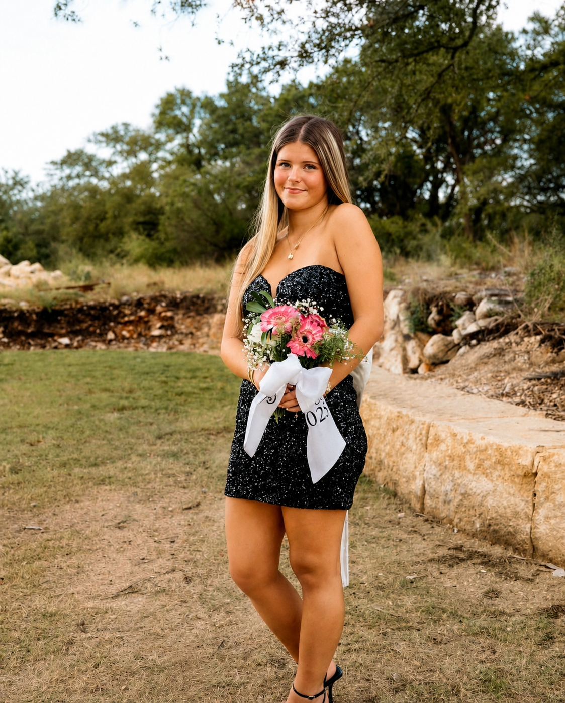 Young woman in black dress holding a bouquet of pink flowers with white ribbon in an outdoor park setting with trees and rocks in the background.
