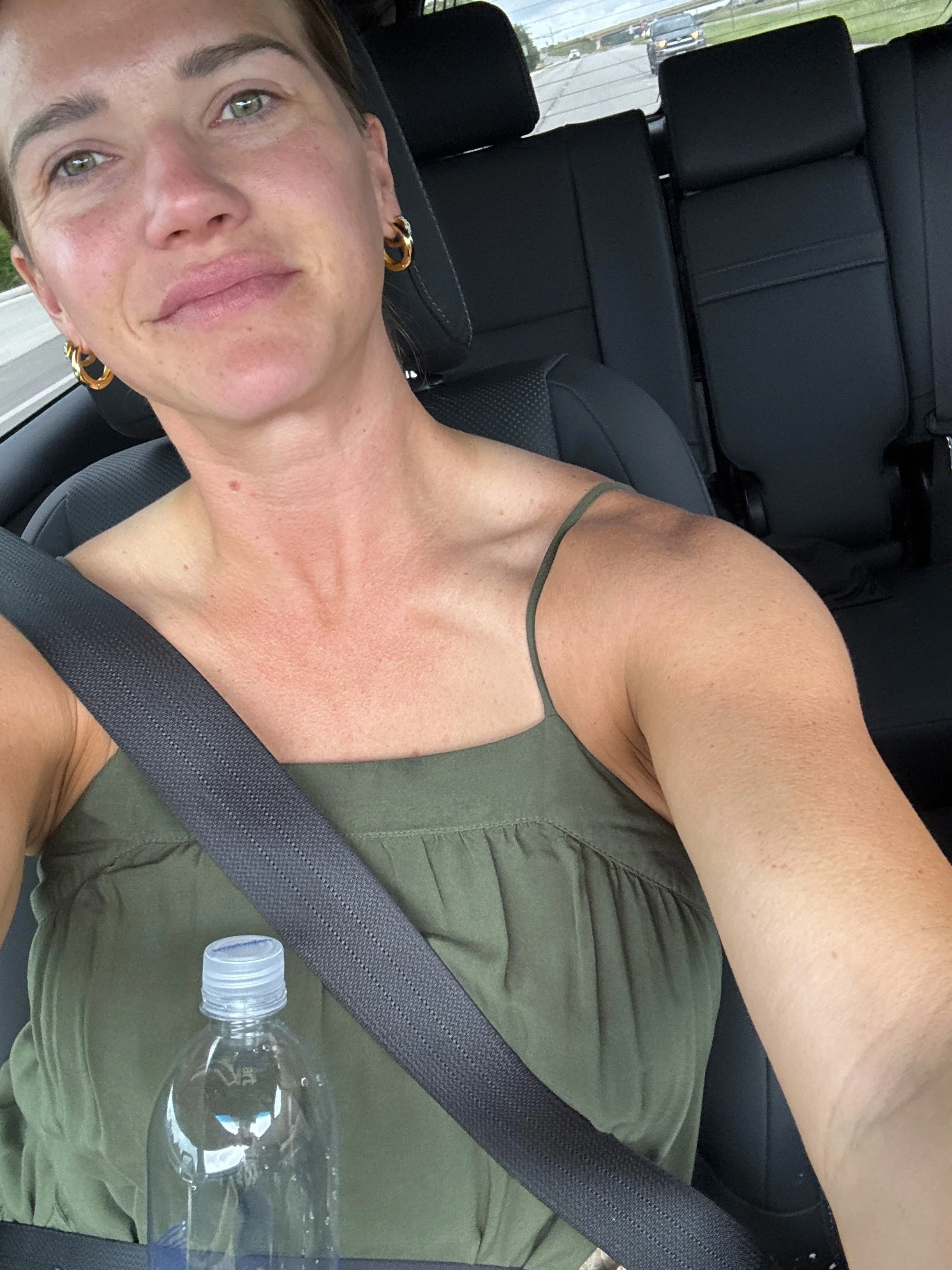 Close-up of a woman with short dark hair, gold hoop earrings, and wearing a green tank top, sitting in the back seat of a car with a black interior, a water bottle on her lap, and other cars visible on the road outside.