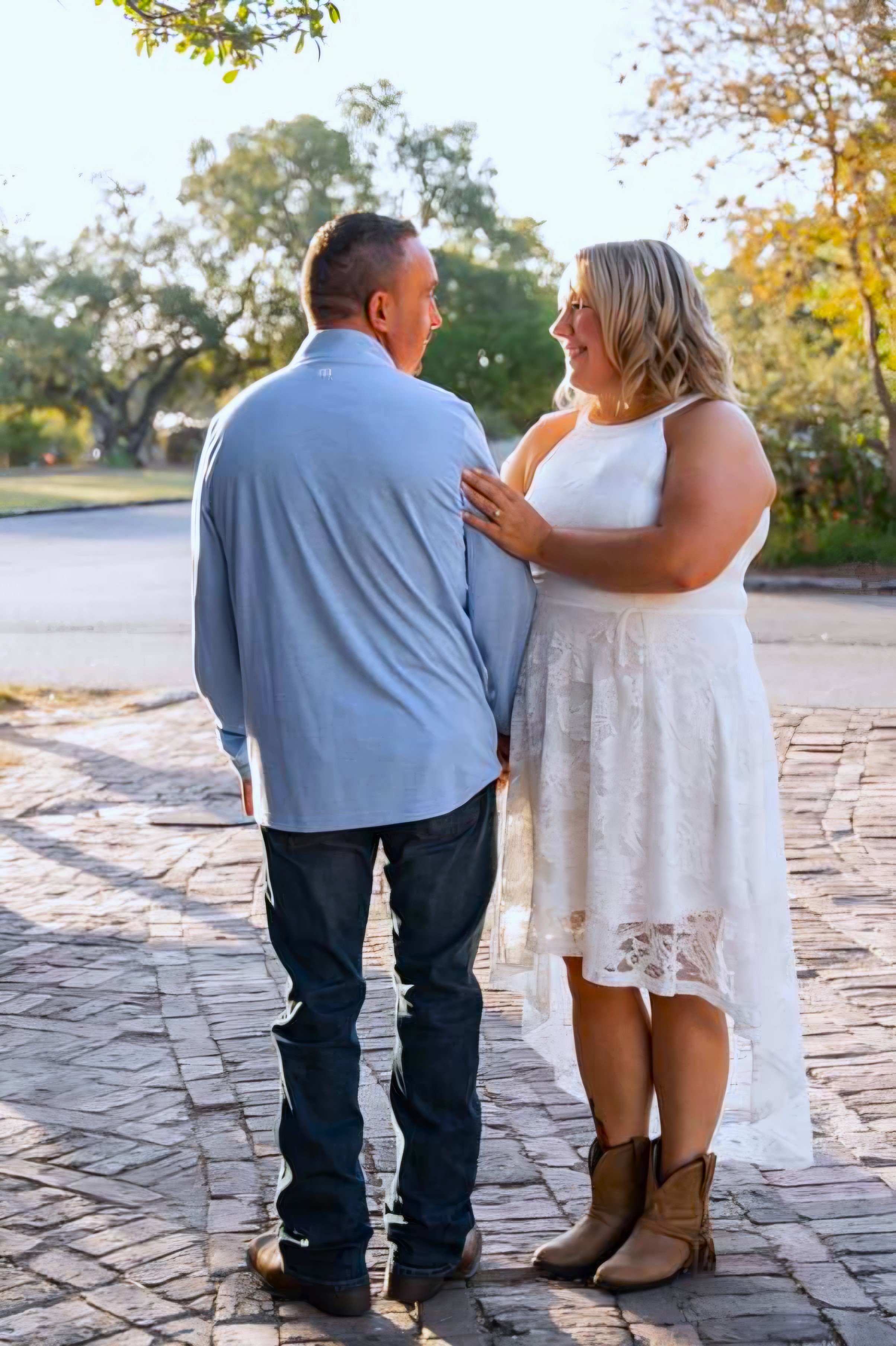 A man and woman standing closely outdoors on a paved path, smiling at each other, with trees and sunlight in the background.