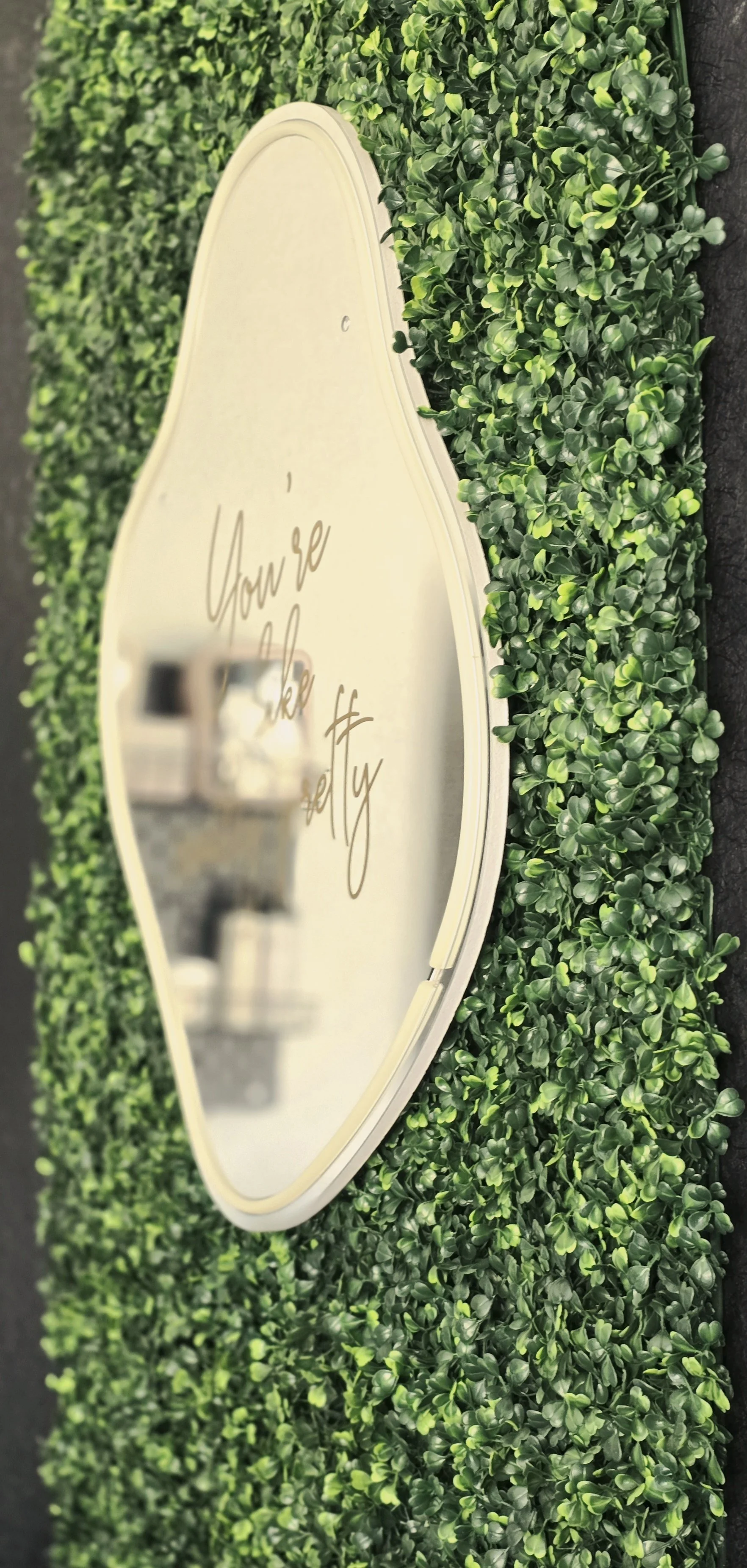 Oval mirror with a white frame hanging on a wall covered with green leaves. The mirror has a quote that says, 'You're like really' in cursive lettering. Reflection of two glasses is visible in the mirror.