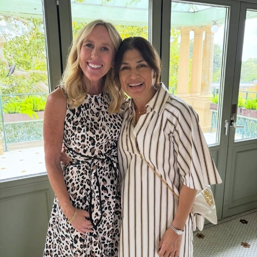 Two women smiling and posing together indoors near large windows with greenery outside.