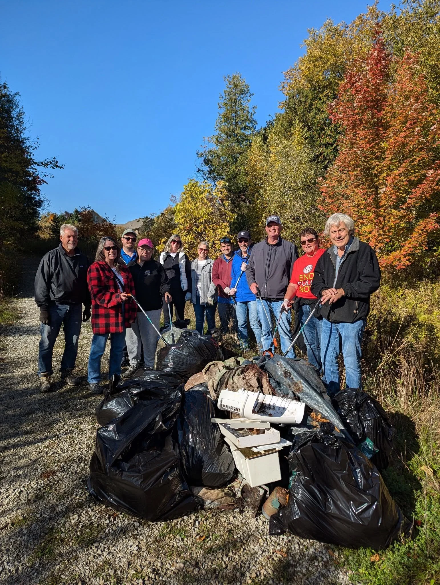 A group of people standing outdoors next to a large pile of trash bags and discarded debris, with trees showing autumn foliage in the background.