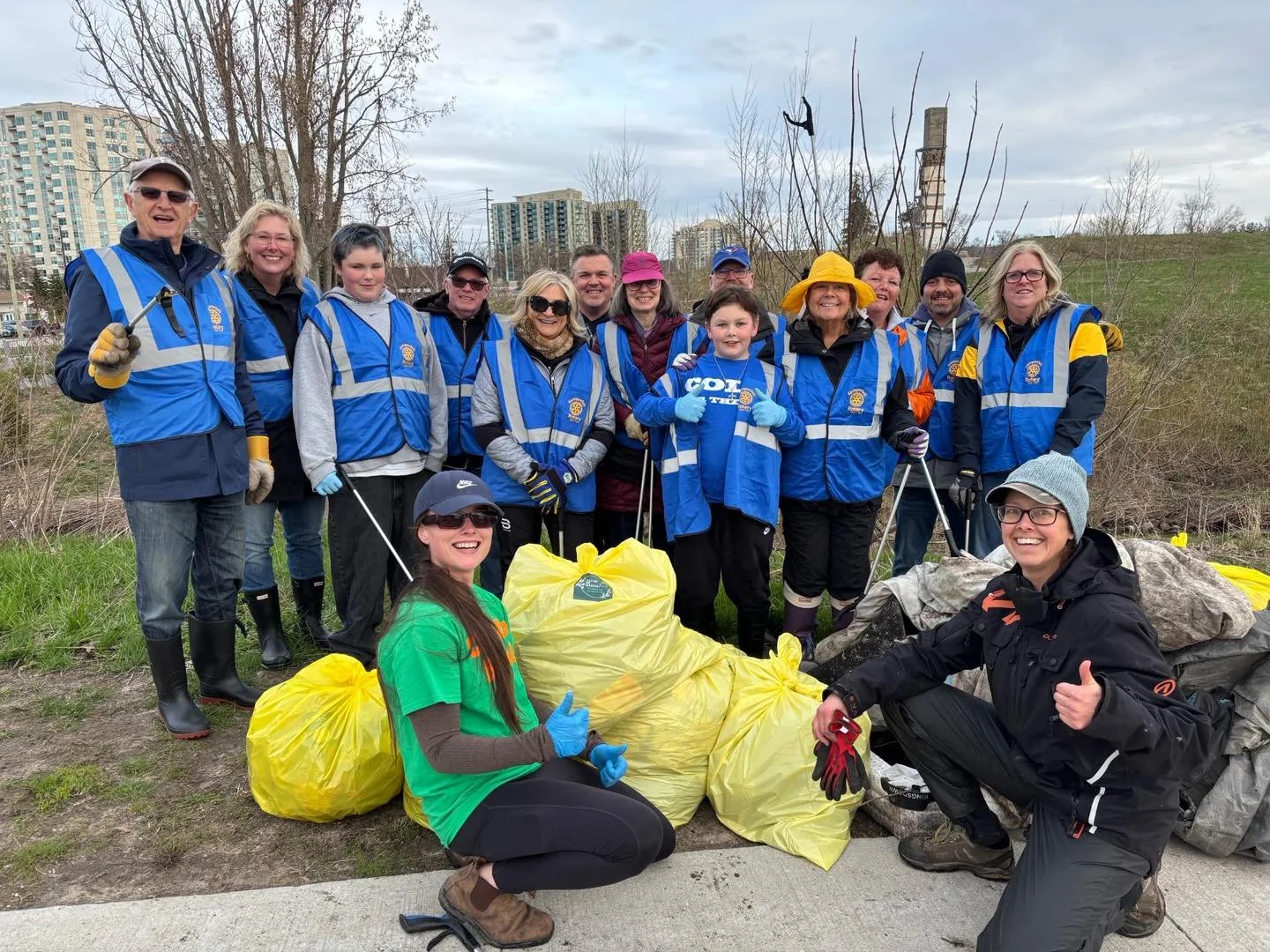 Group of people wearing blue volunteer vests and gloves, participating in a cleanup event outdoors with large yellow trash bags filled with collected litter, with a cityscape in the background.