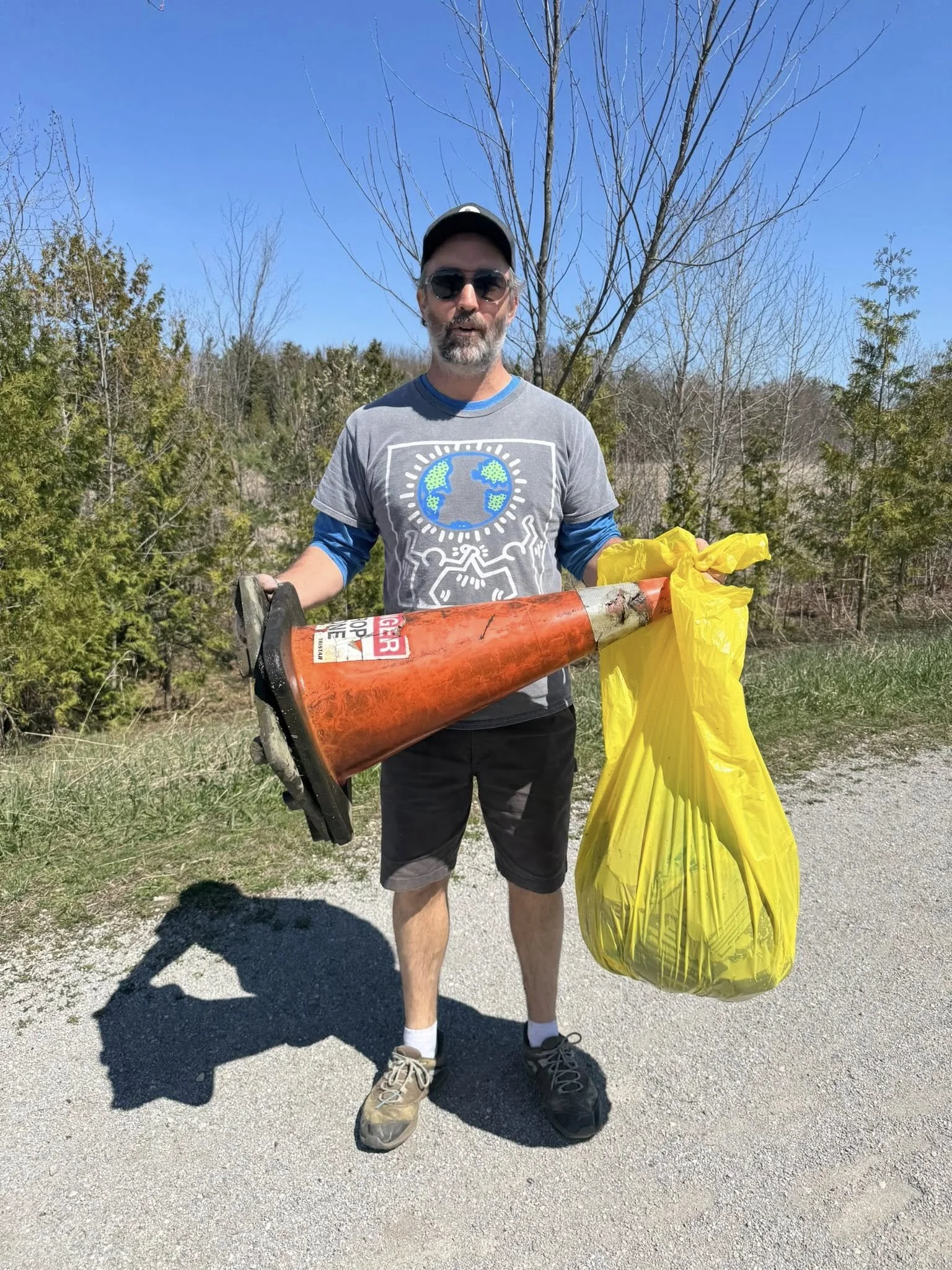 A man wearing sunglasses and a gray t-shirt with a globe and earth design stands outdoors on a sunny day, holding a large orange fire extinguisher and a yellow bag, with trees and blue sky in the background.