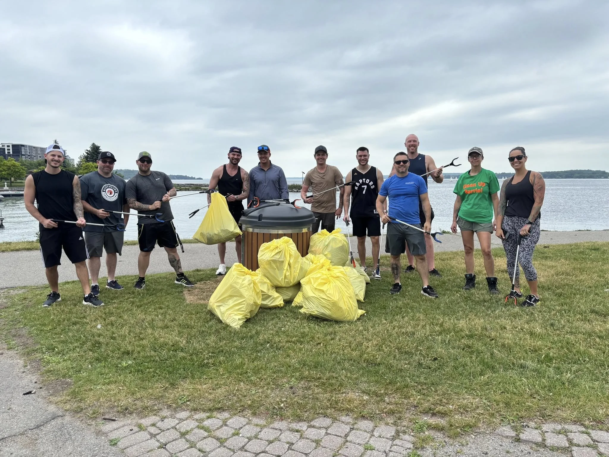Group of eleven people standing by a body of water, holding trash pickers, with yellow trash bags filled with collected litter, during daytime with overcast sky.