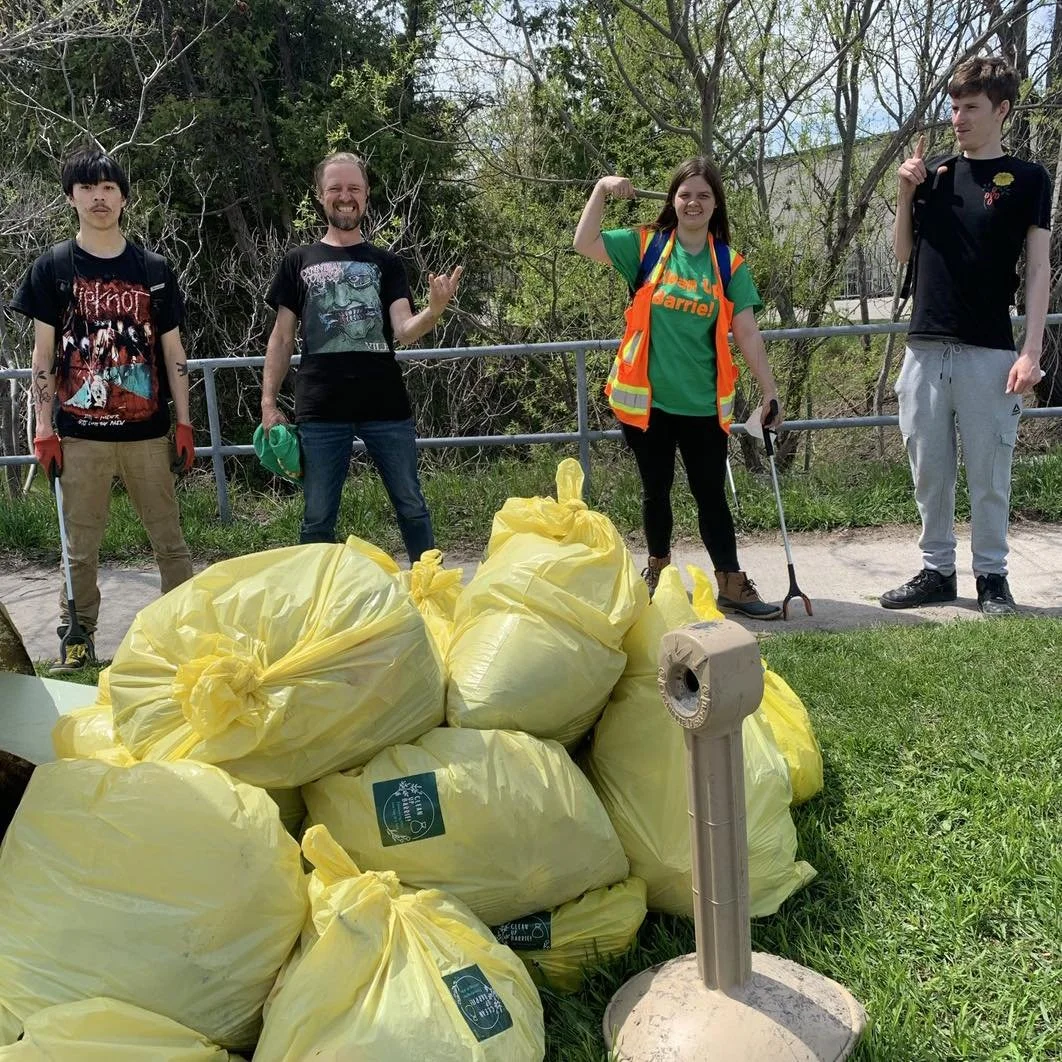 Four people standing outdoors behind a pile of yellow trash bags filled with litter. They are smiling and posing, wearing casual clothing and gloves, with tools for cleanup. One person is wearing a high-visibility vest. The background has trees and a metal fence.