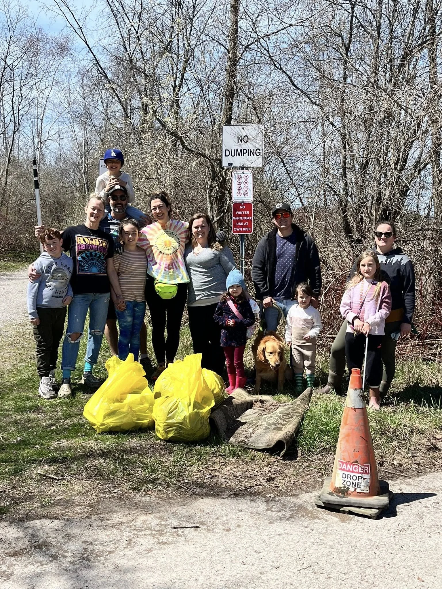 A group of people, including children and adults, standing outdoors in front of a wooded area with leafless trees, holding trash bags and trash pick-up tools. There is a 'No Dumping' sign and an orange cones with a 'Danger Drop Zone' sign on it in the foreground, and yellow trash bags and a rolled-up carpet on the ground.