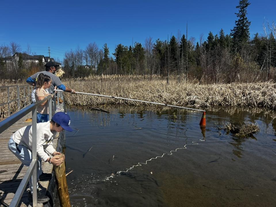 Children and adults fishing in a river from a wooden dock, with some using a fishing pole and others preparing to fish, in a natural outdoor setting with trees and blue sky.