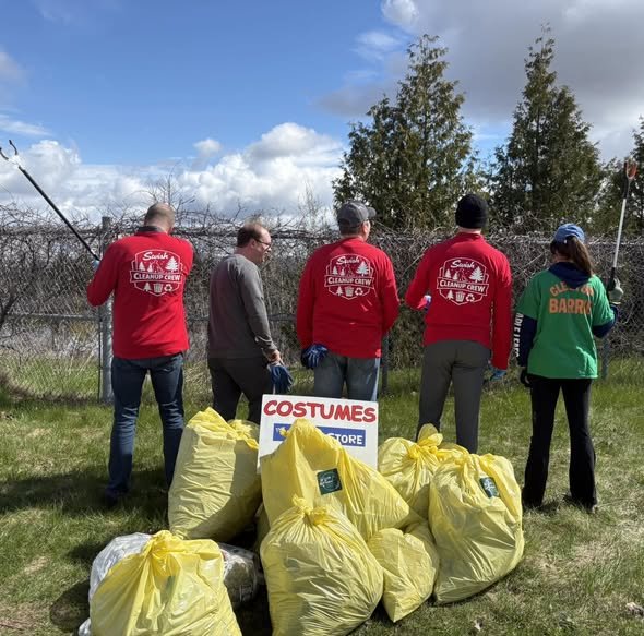 Group of five people wearing red and green shirts participating in a community cleanup event, standing on grass near a chain-link fence with trees in the background, yellow trash bags filled with collected litter in the foreground, a sign that reads 'COSTUMES' with a store logo.