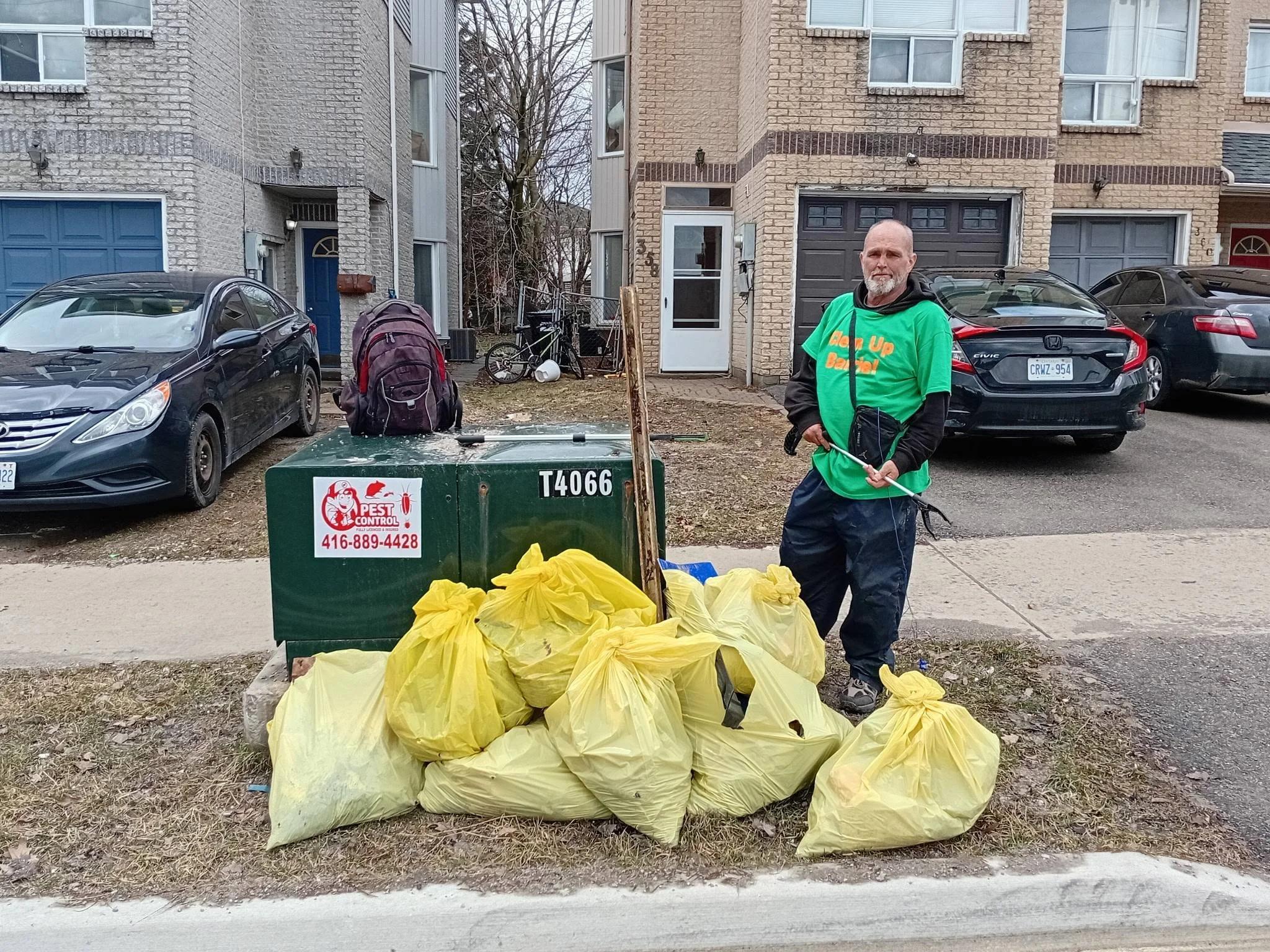 A man wearing a green shirt and black pants holding a stick next to a large green trash bin overflowing with yellow garbage bags on a residential street.