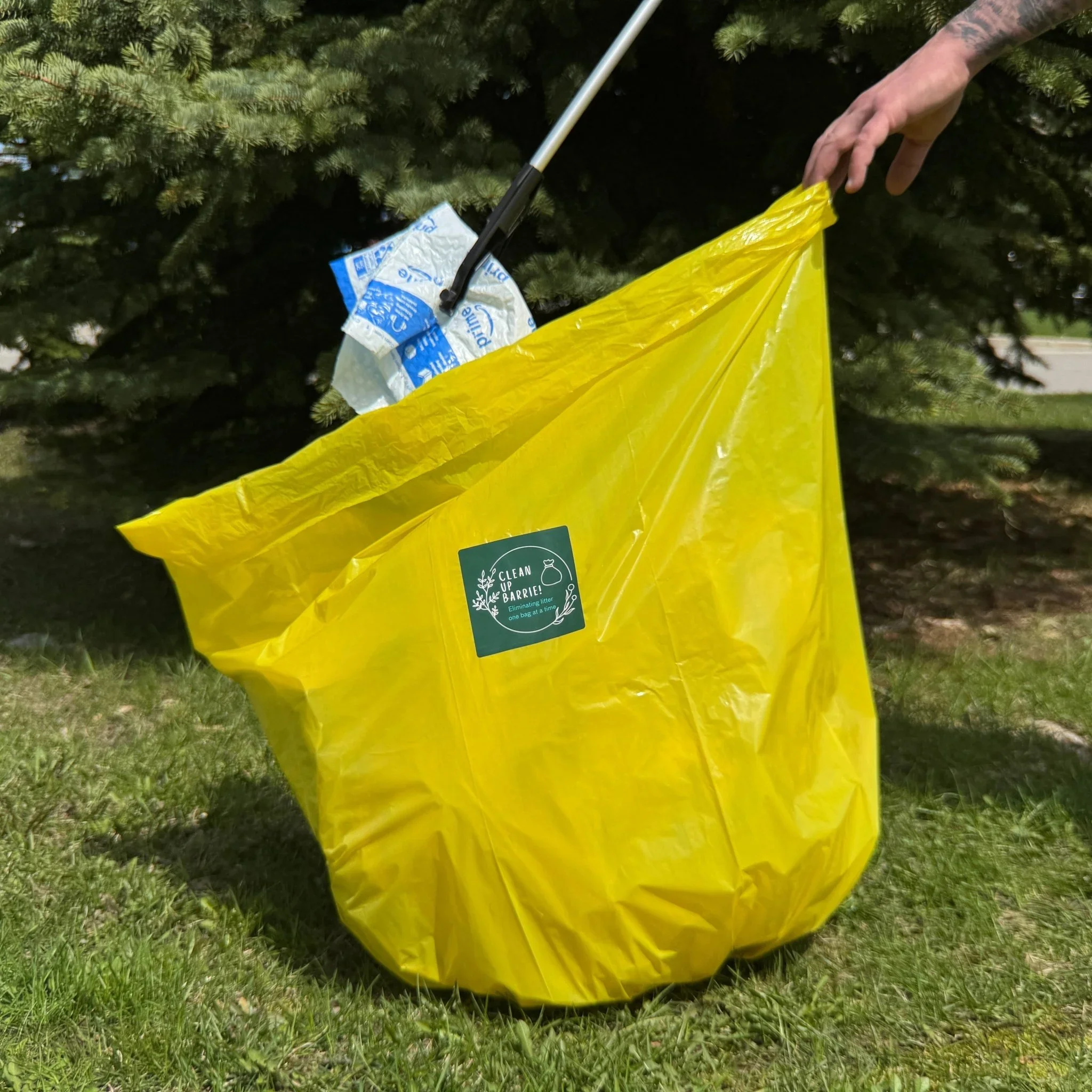 A yellow trash bag with a green label on grass, being filled with litter from a person’s hand.