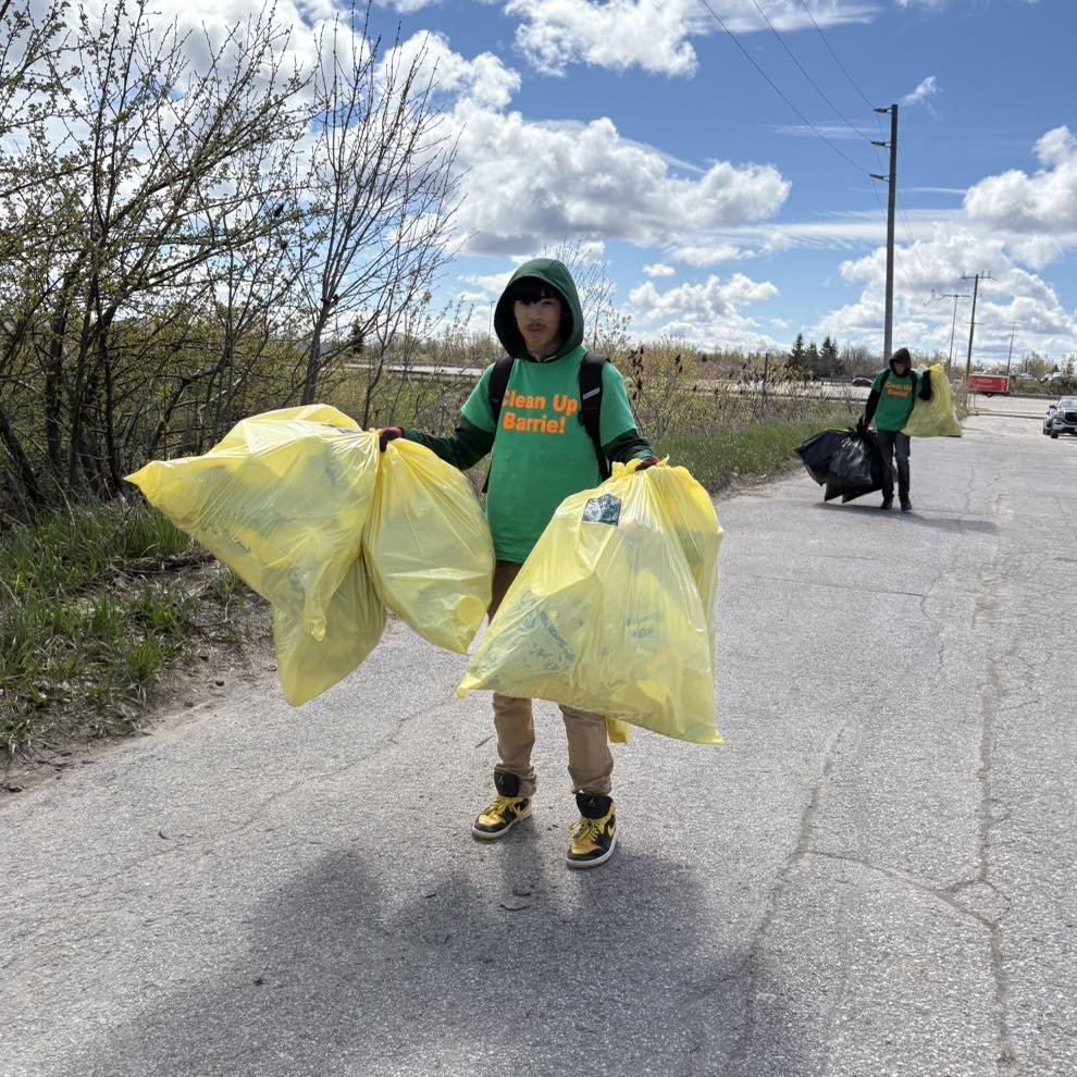 Two young volunteers picking up trash along the roadside, wearing green shirts and holding large yellow trash bags. The sky is partly cloudy, and the area shows some sparse trees and roadside infrastructure.