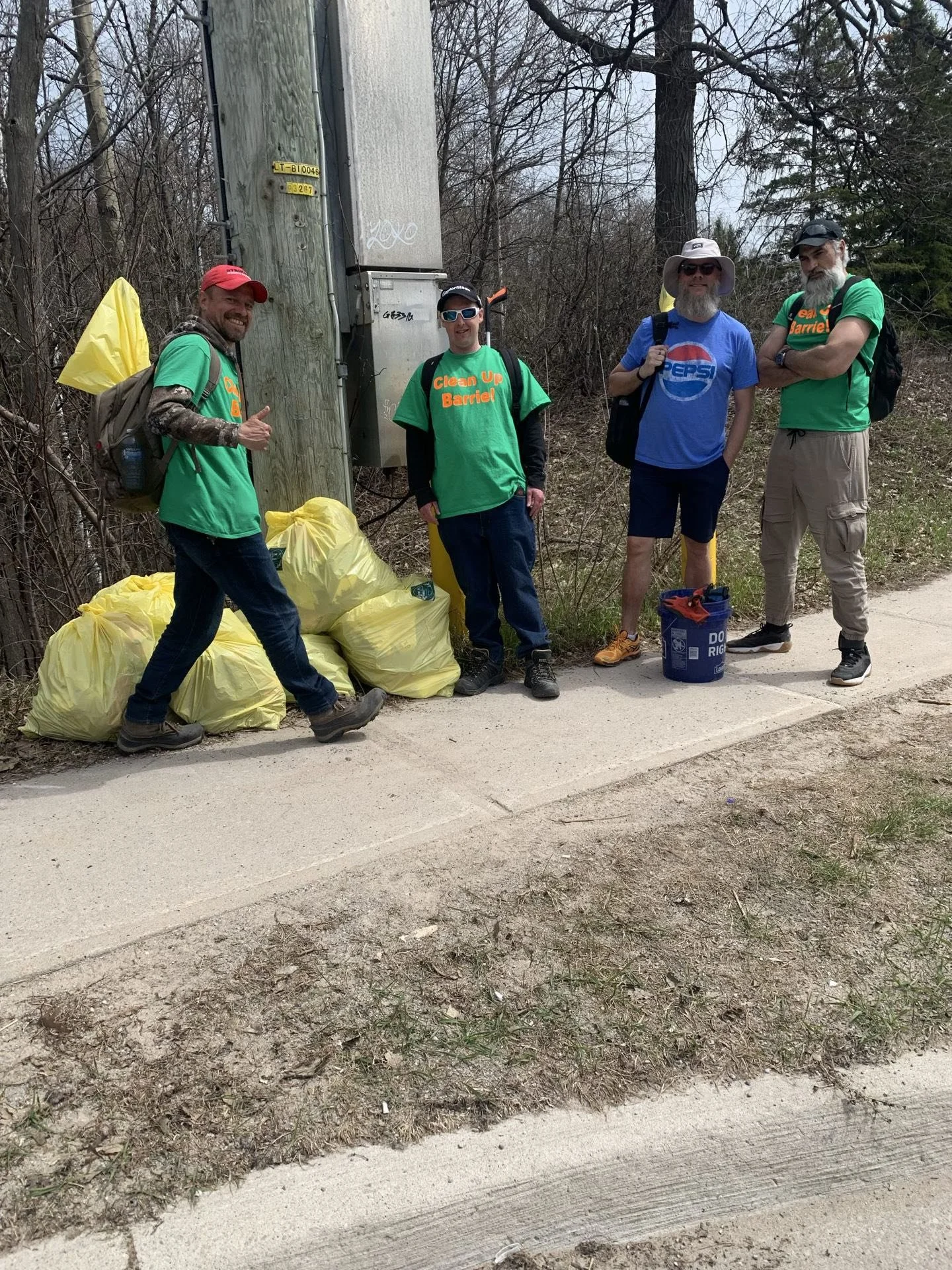 Four men participating in a community cleanup event on a sidewalk with trees and bushes in the background. They are standing beside yellow trash bags filled with collected litter. The men are wearing green shirts that read "Clean Up Barrel!" and are equipped with backpacks, hats, and sunglasses.