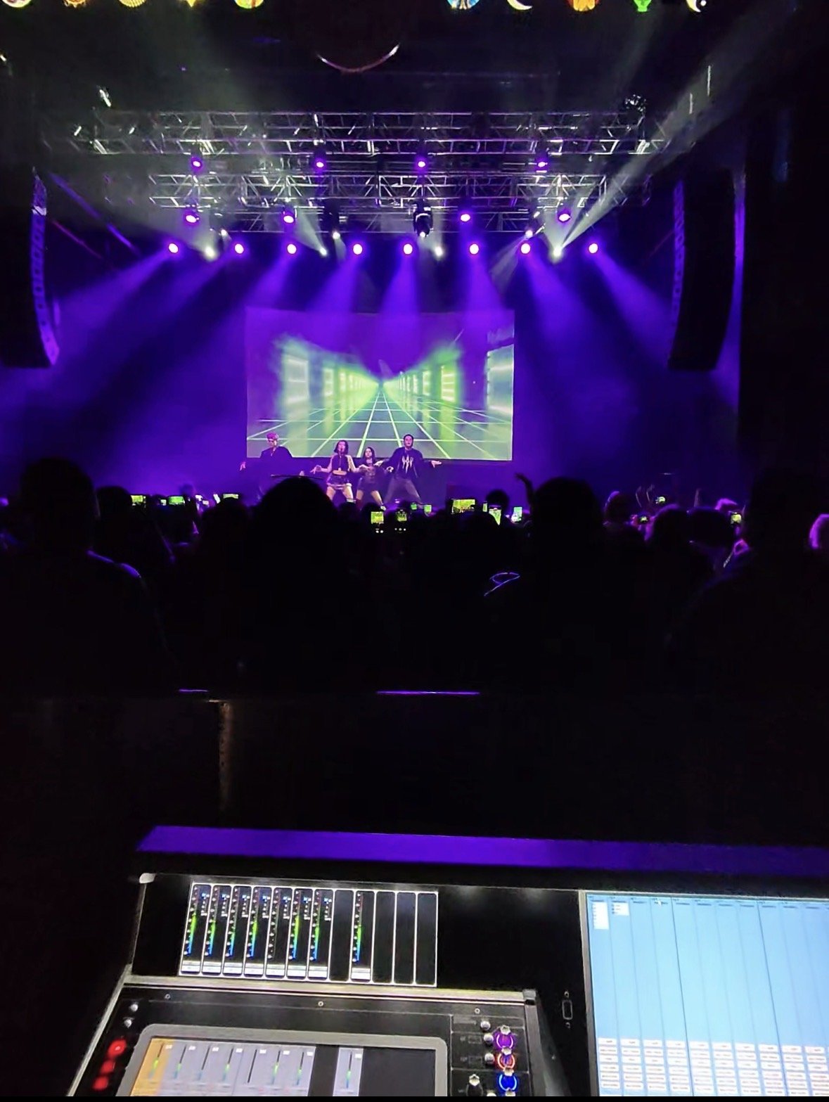 A stage with performers and vibrant purple lighting, featuring a digital backdrop with a futuristic grid and tunnel design. The audience is visible, and a sound mixing console is in the foreground.